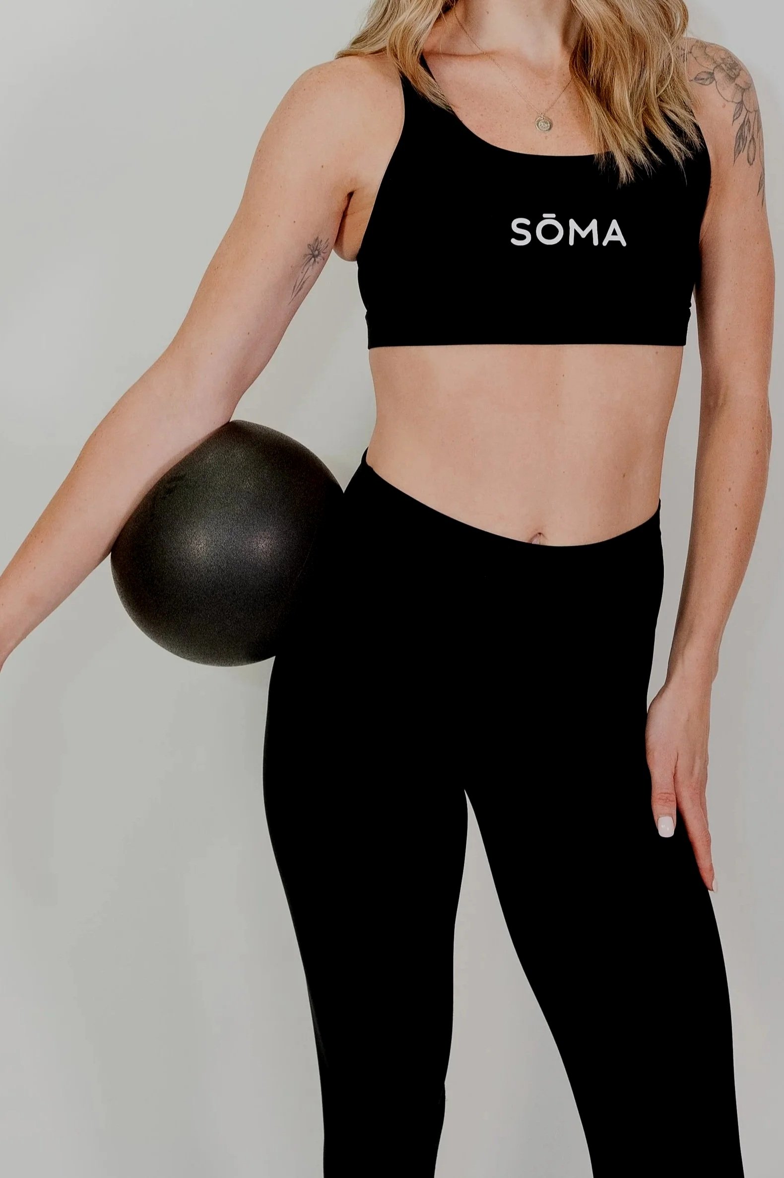 A woman wearing a black sports bra with the word 'SÖMA' printed on it, holding a black exercise ball, dressed in black leggings, facing slightly to the right, with tattoos on her shoulders and arms, standing against a plain white background.