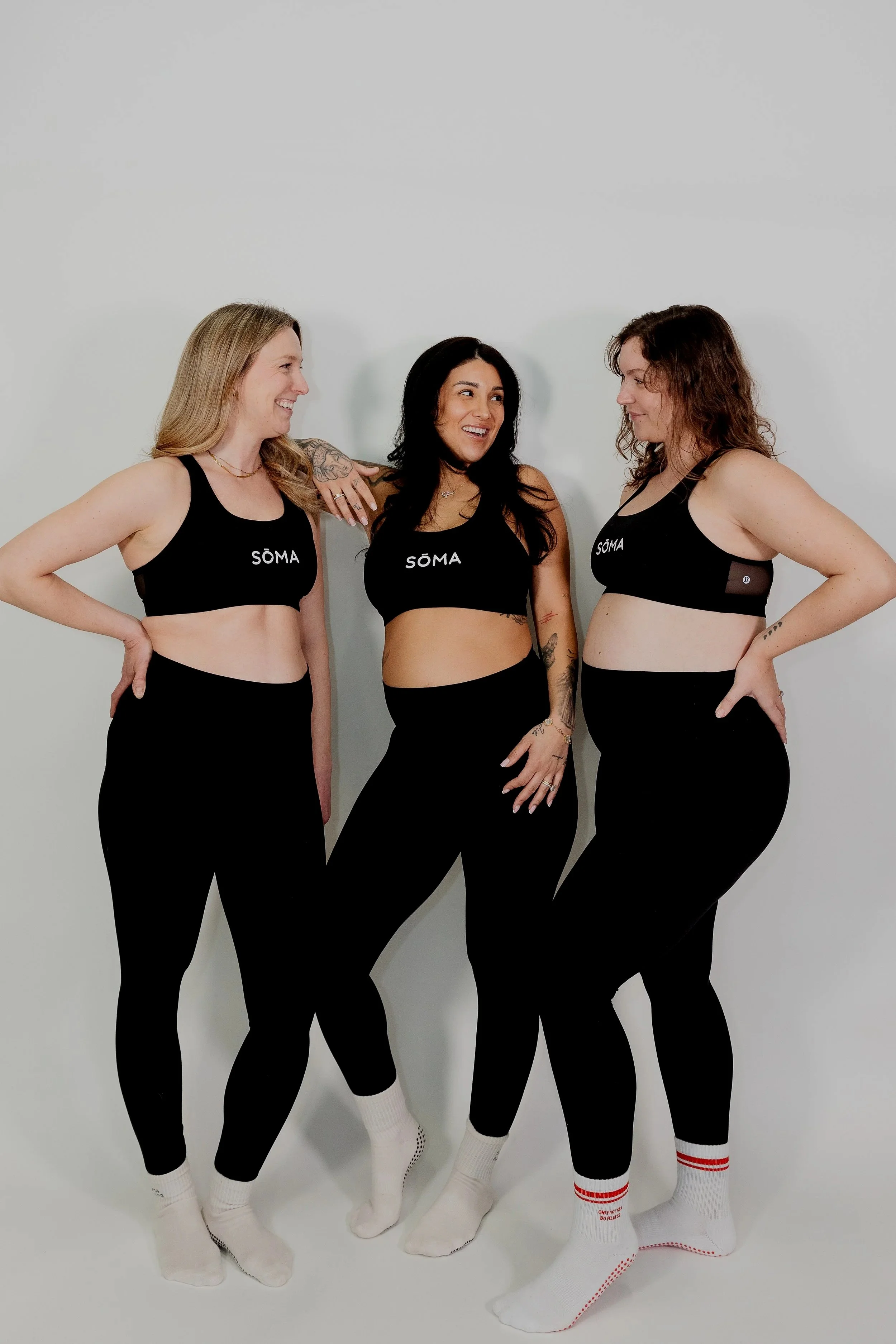 Three women standing together against a white background, wearing matching black sports bras with the word 'SÖMA' printed on them and black leggings, smiling and engaging in conversation.