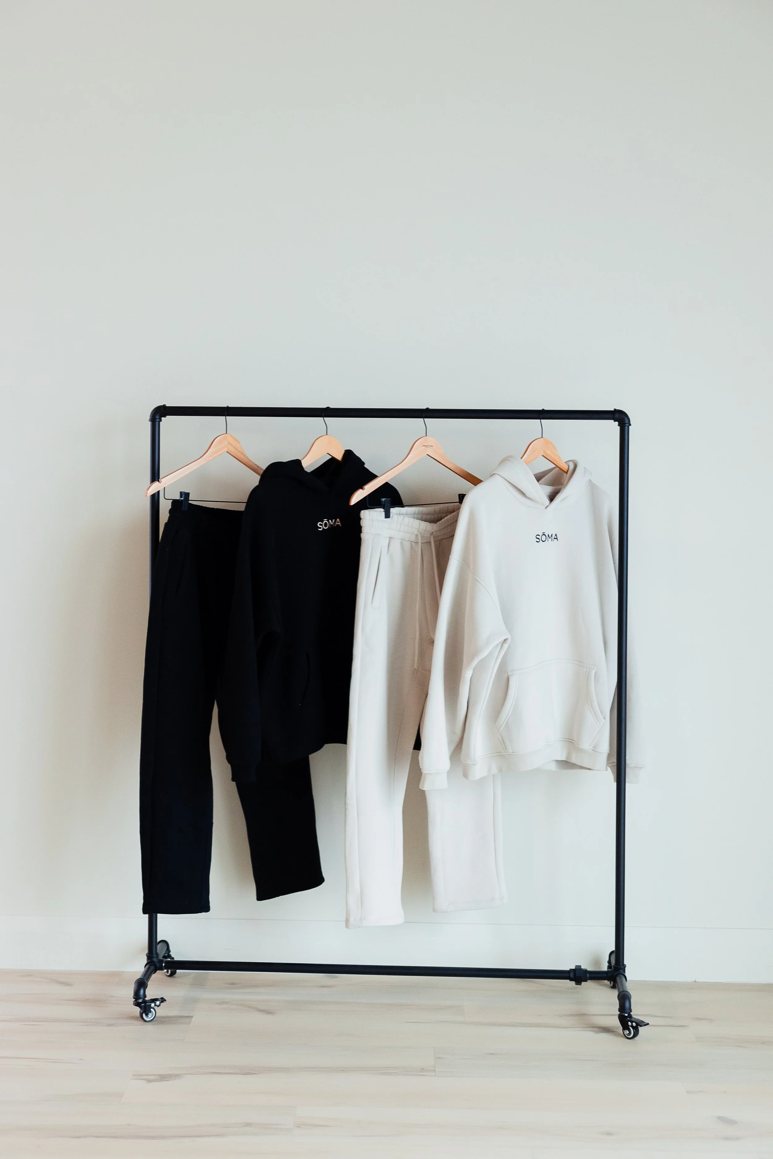 Clothing rack with black and white hoodies and pants on wooden hangers against a plain wall.