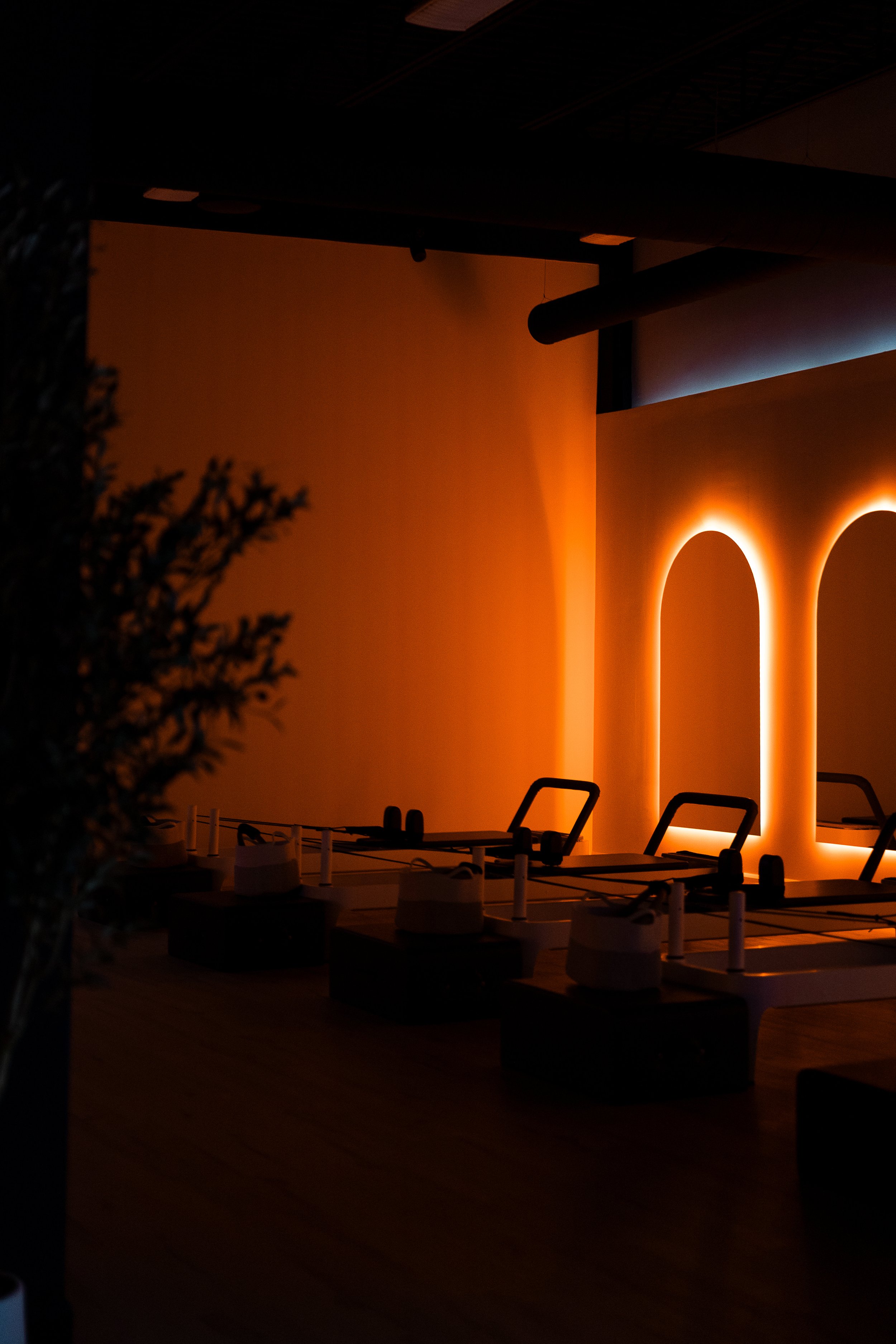 Interior of a  pilates studio with warm orange lighting, modern design, and empty tables with chairs. There are illuminated arches on the wall and some plants in the foreground.