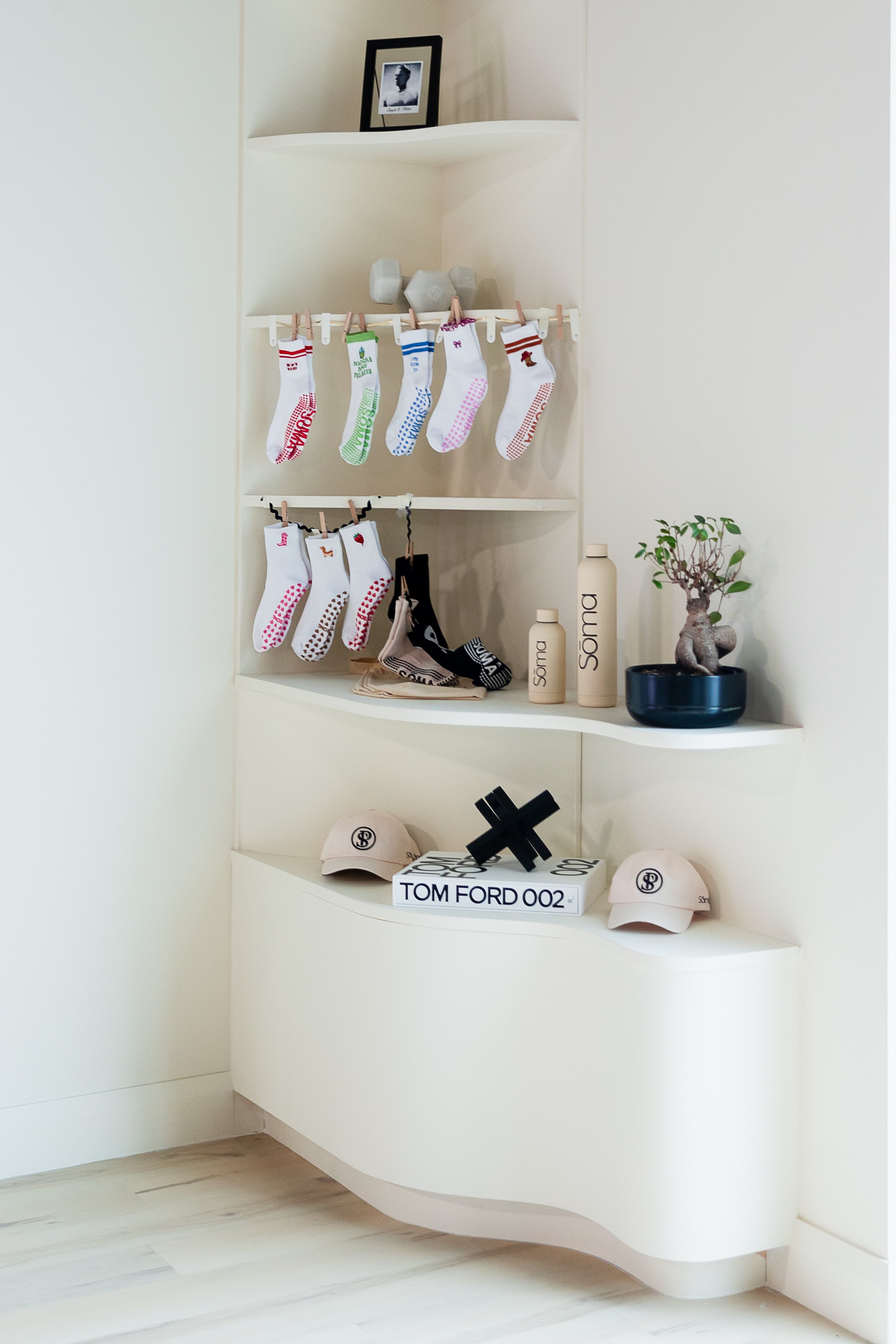 Decorative shelf with grip socks, beige hats, and bottles labeled 'Soma', topped with a small bonsai tree in a black pot, and a black decorative object, in a minimalist room with light-colored wooden flooring.
