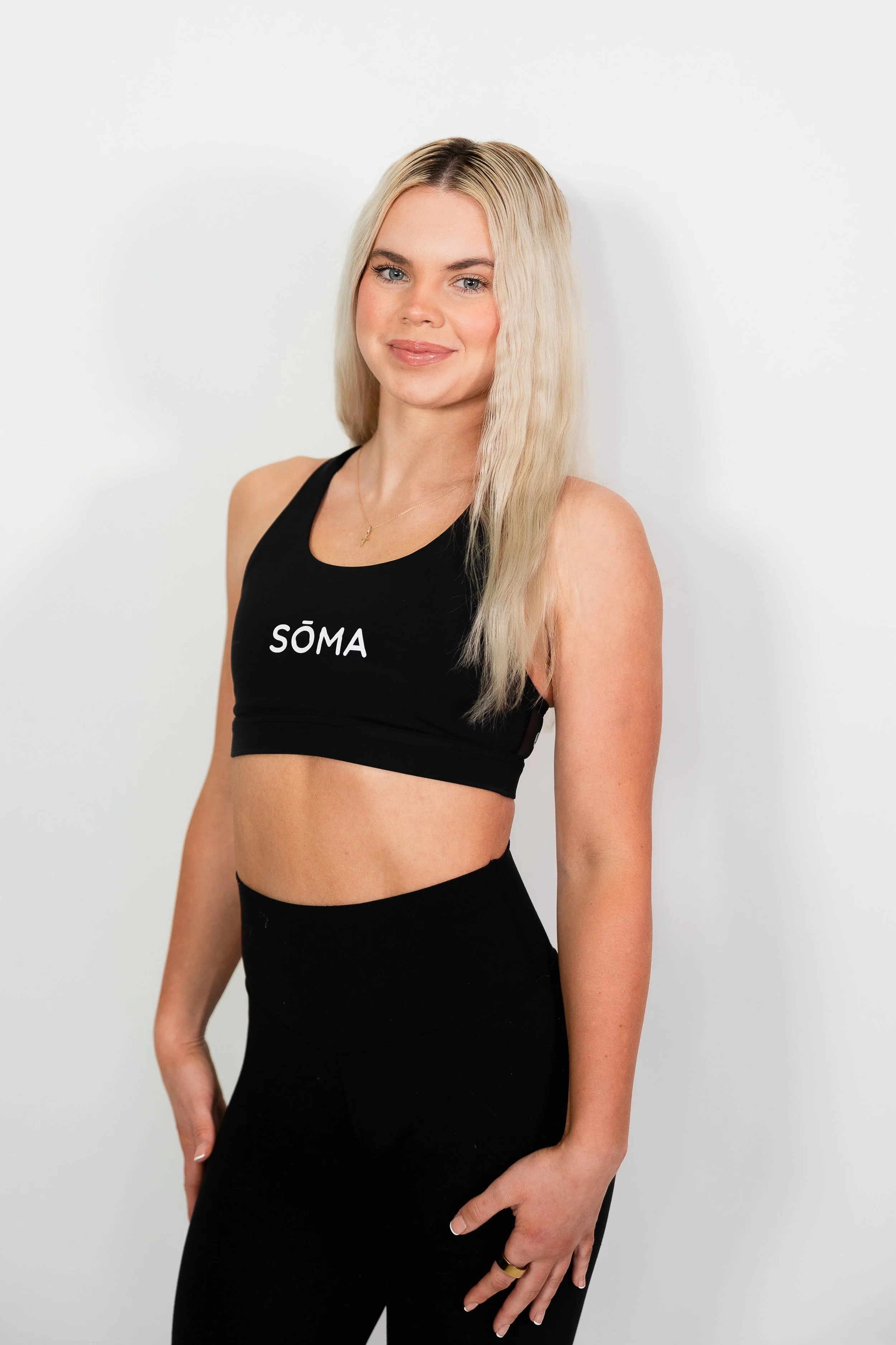 A young woman with blonde hair wearing a black sports bra with the word 'SÖMA' on it and black leggings, standing against a plain white background.