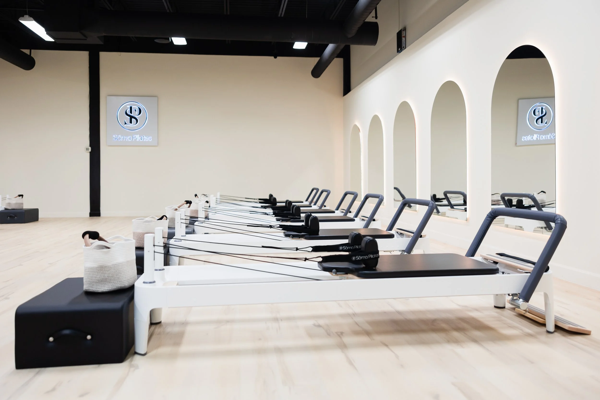 Indoor Pilates studio with reformer equipment lined up, illuminated mirrors on the right wall, and a modern design with black and white accents.