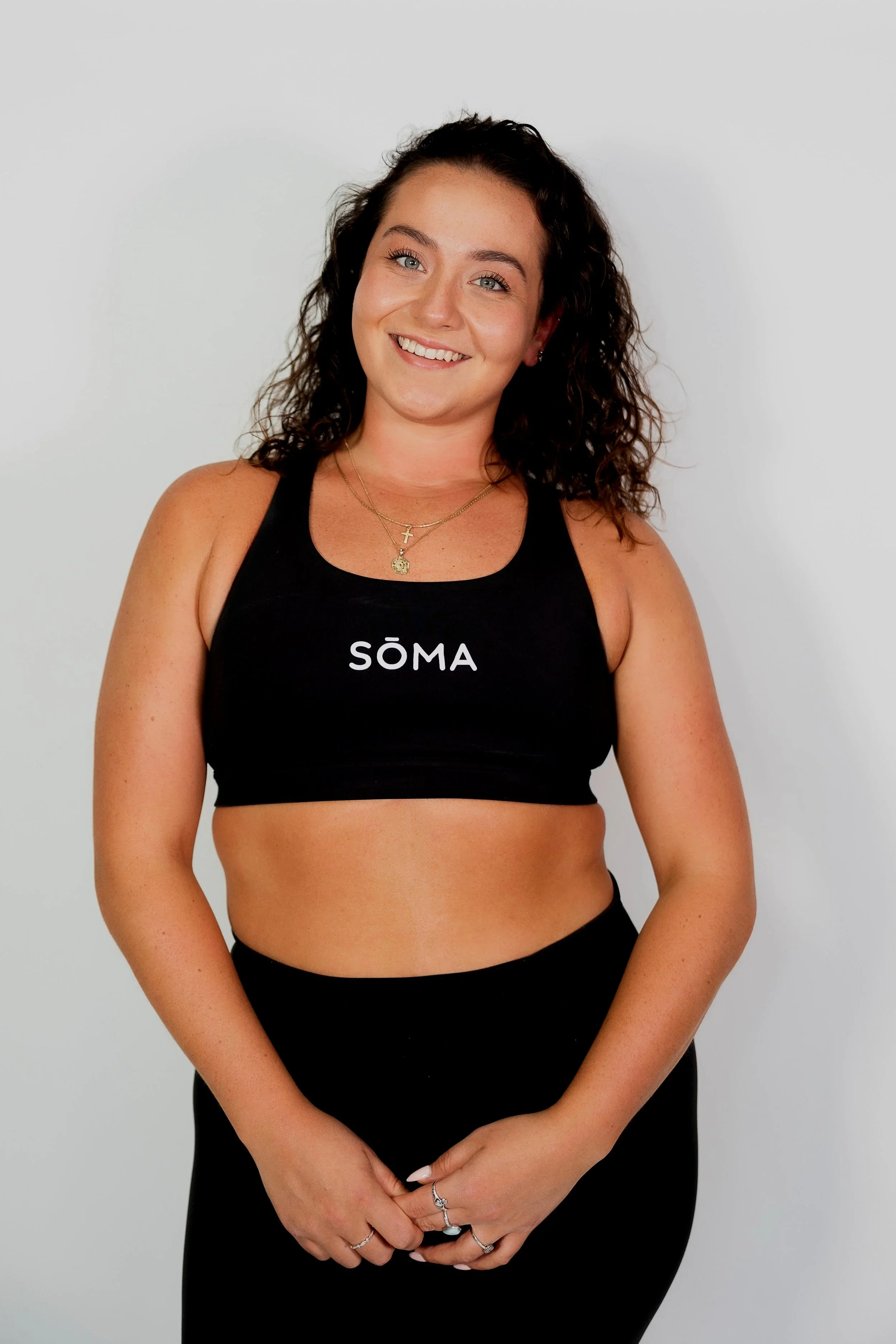 Smiling woman with curly brown hair wearing a black sports bra with 'SÖMA' written on it, standing against a plain white background.
