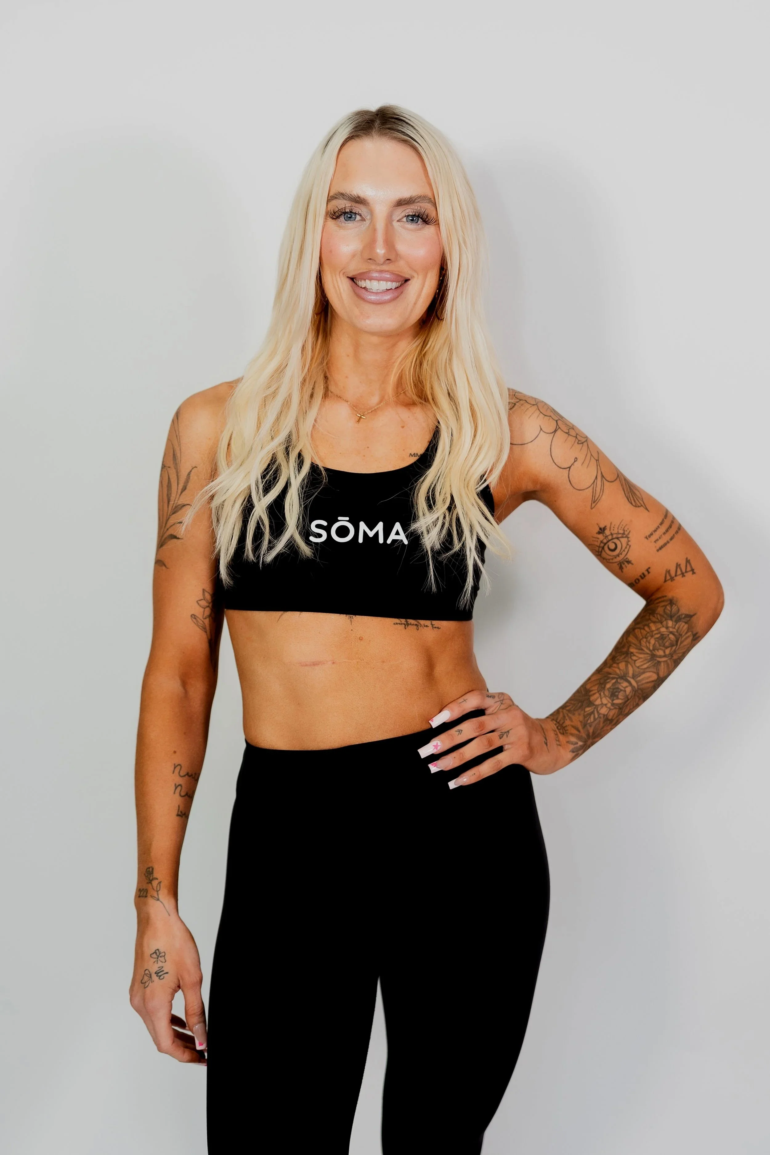 A woman with long blonde hair, tattoos on her arms, wearing a black athletic top with the word 'SÖMA' on it, and black pants, standing against a plain white background, smiling and looking at the camera.