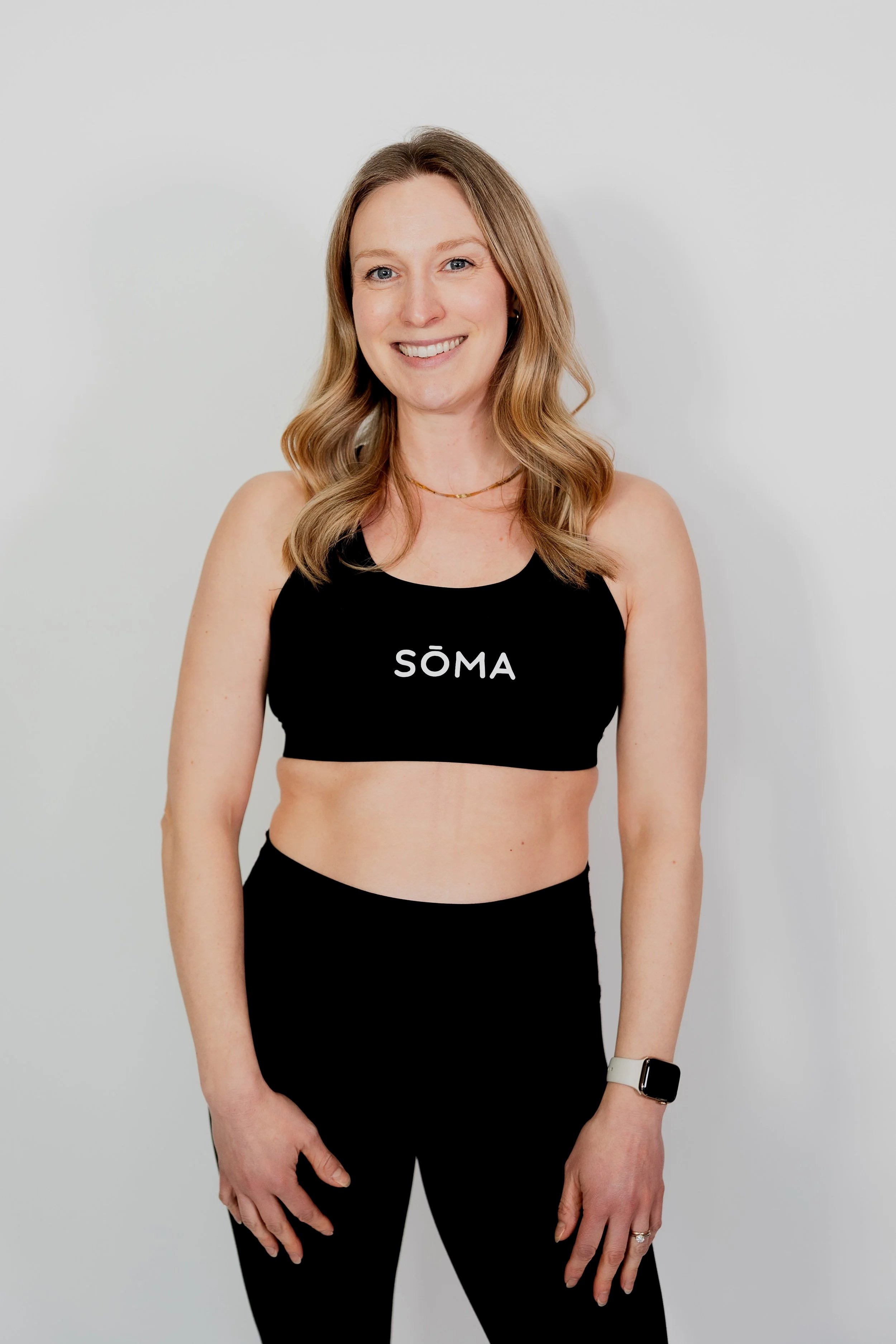A smiling woman with long wavy hair wearing a black sports bra with the word 'SÖMA' printed on it, black leggings, a smartwatch, and a ring, standing against a plain light gray background.