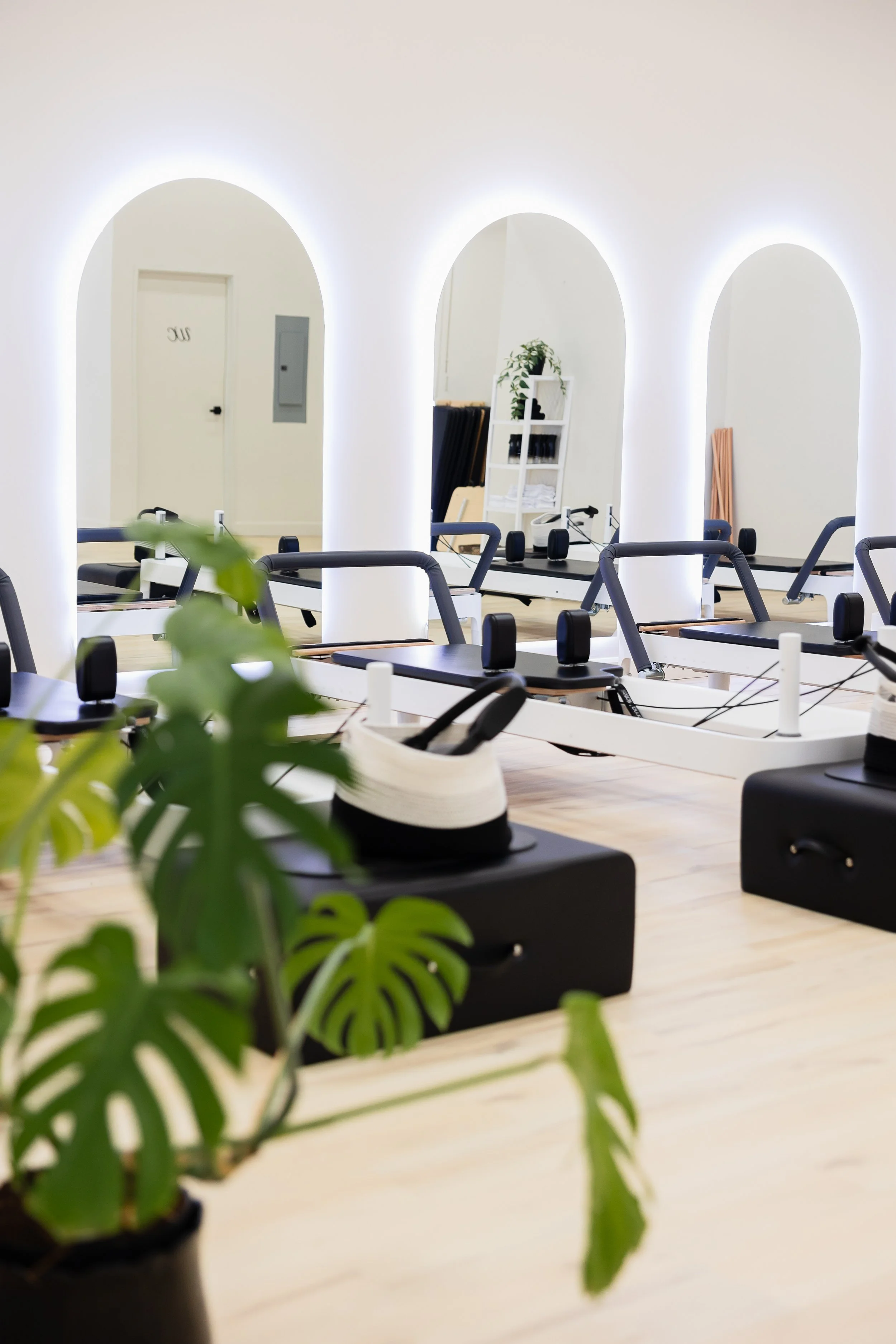 Interior of a modern fitness or Pilates studio with workout equipment and a large mirror, illuminated by white backlighting, with a potted plant in the foreground.