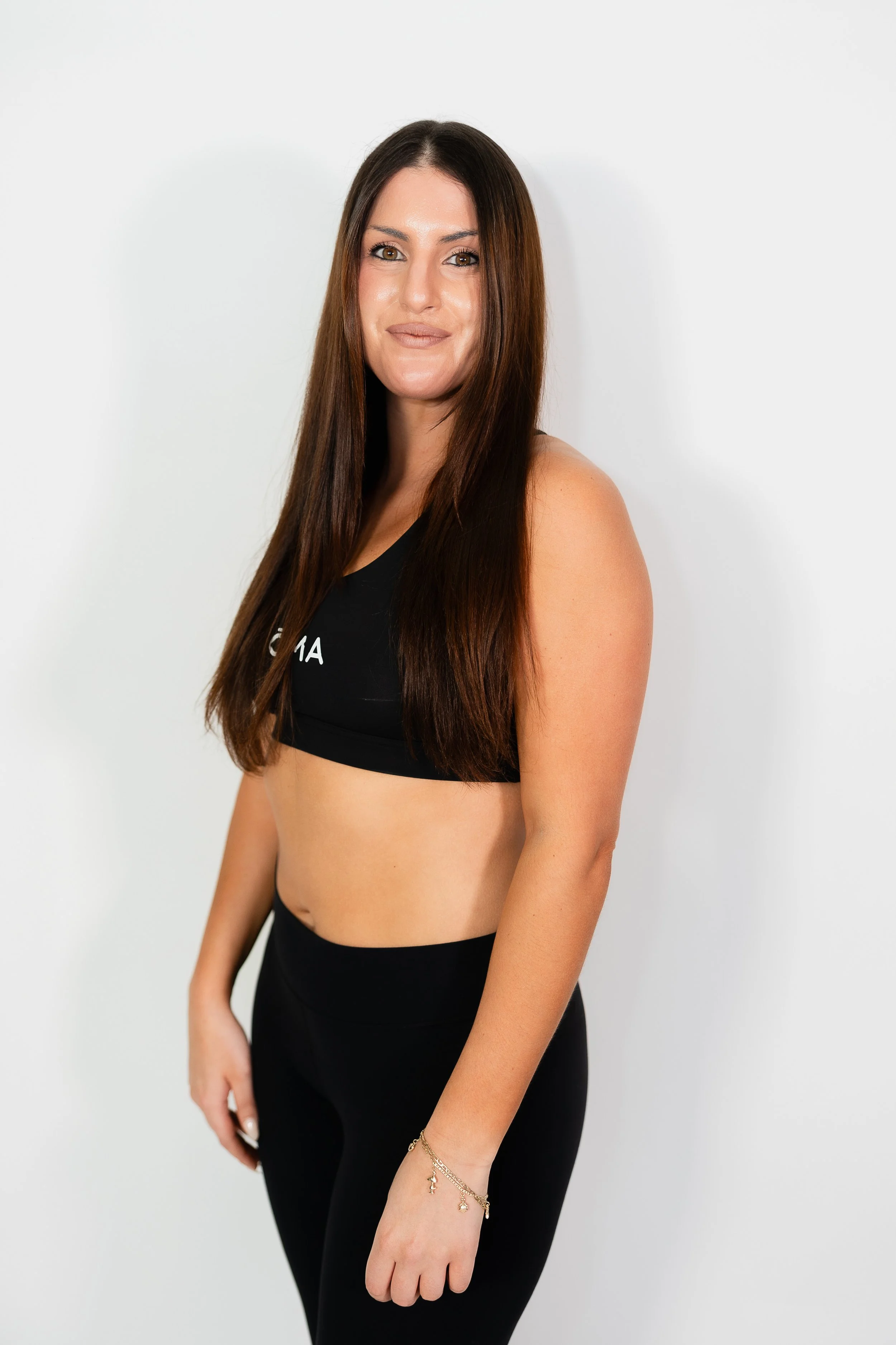 A woman with long brown hair and tan skin wearing a black sports bra with text and black leggings, standing against a plain white wall.