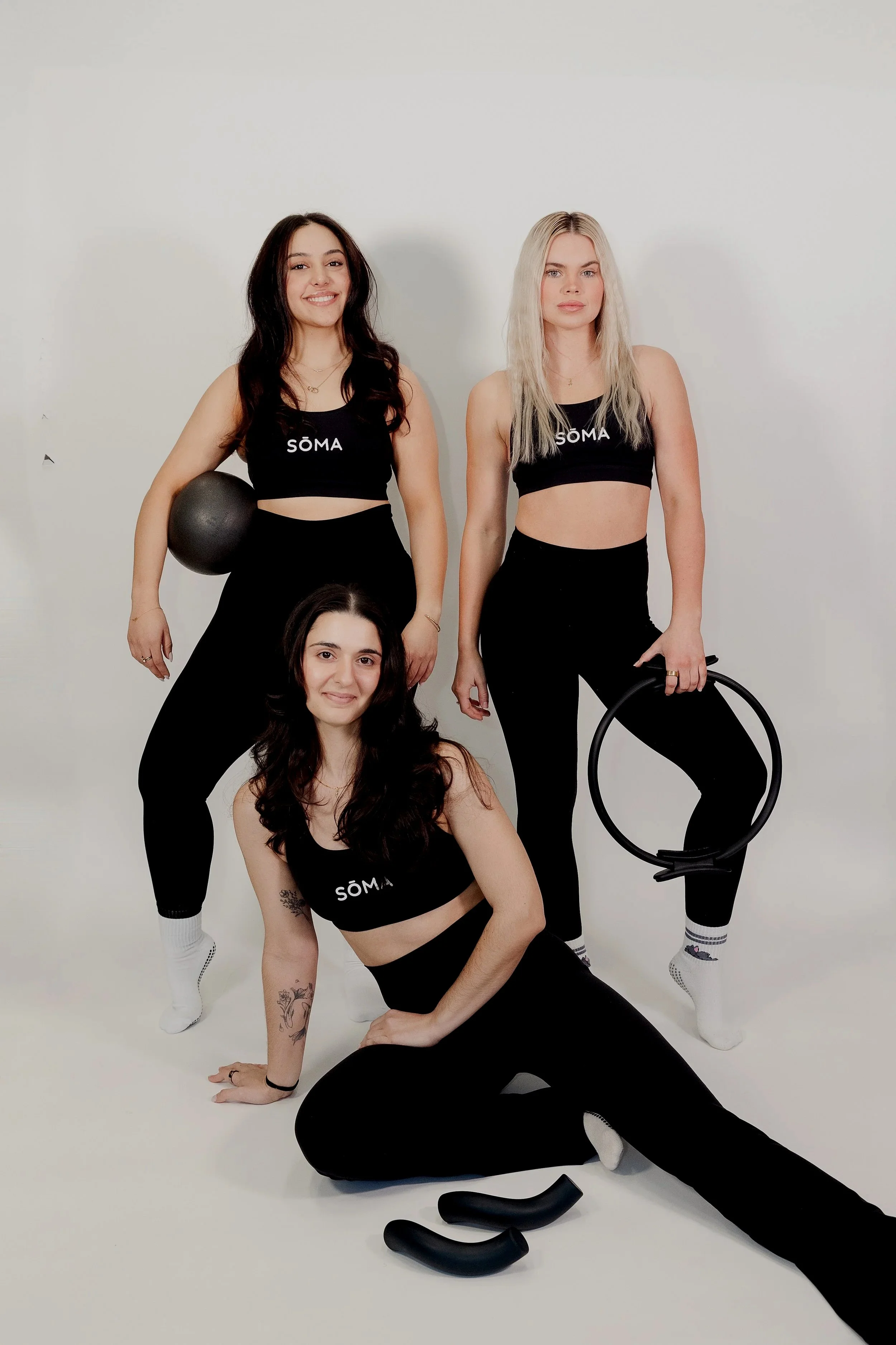 Four women in black workout clothes with the word 'SÖMA' on their tops, posing in front of a white background. One woman is holding a black exercise ball, another is holding a hoop, and one has a pair of black handles on the floor. They are smiling and looking at the camera.