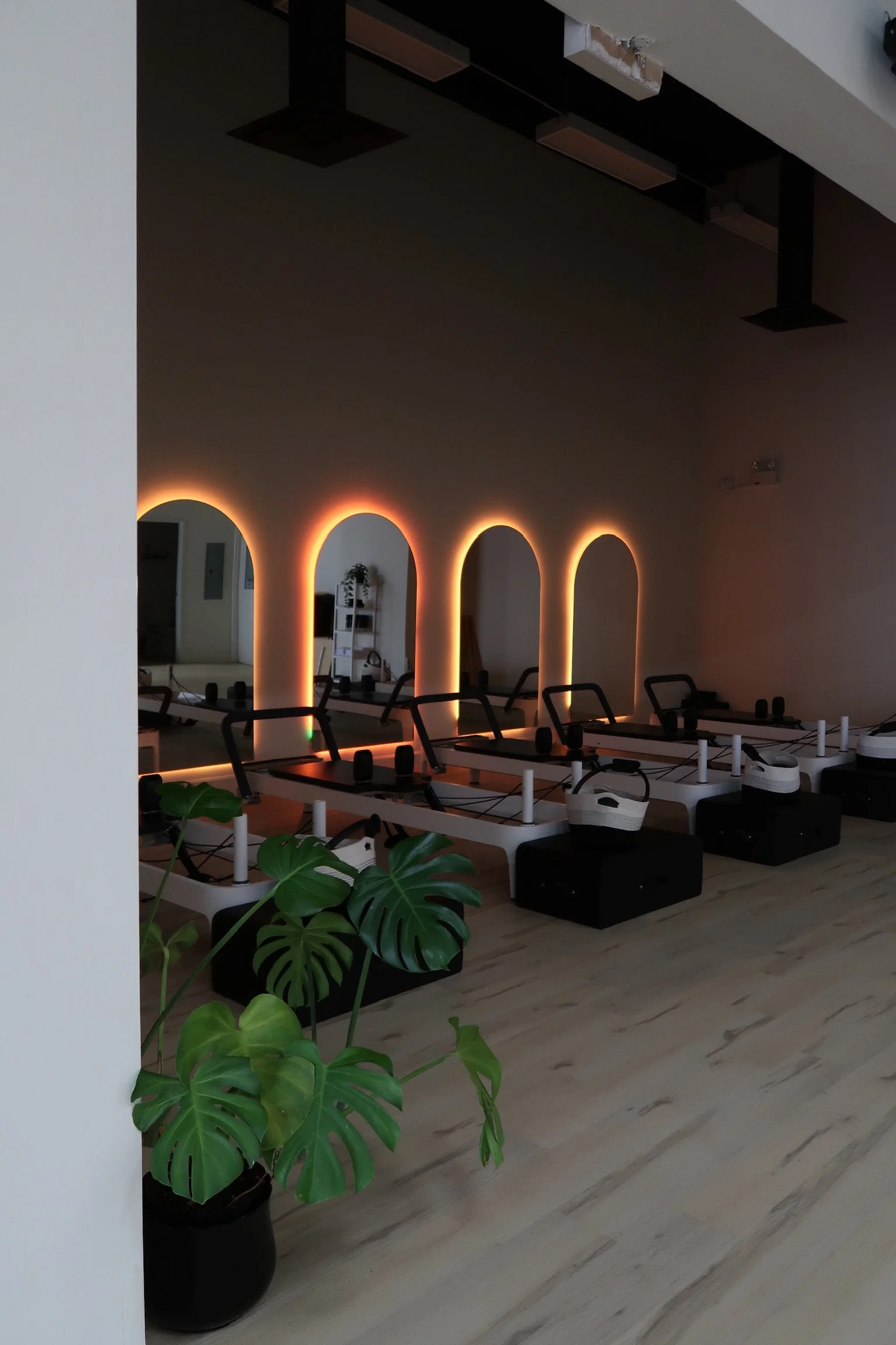 Pilates studio in ottawa with black and white Pilates reformer machines, decorative arches with warm backlighting on the wall, potted plants in black containers, and light-colored wood flooring.