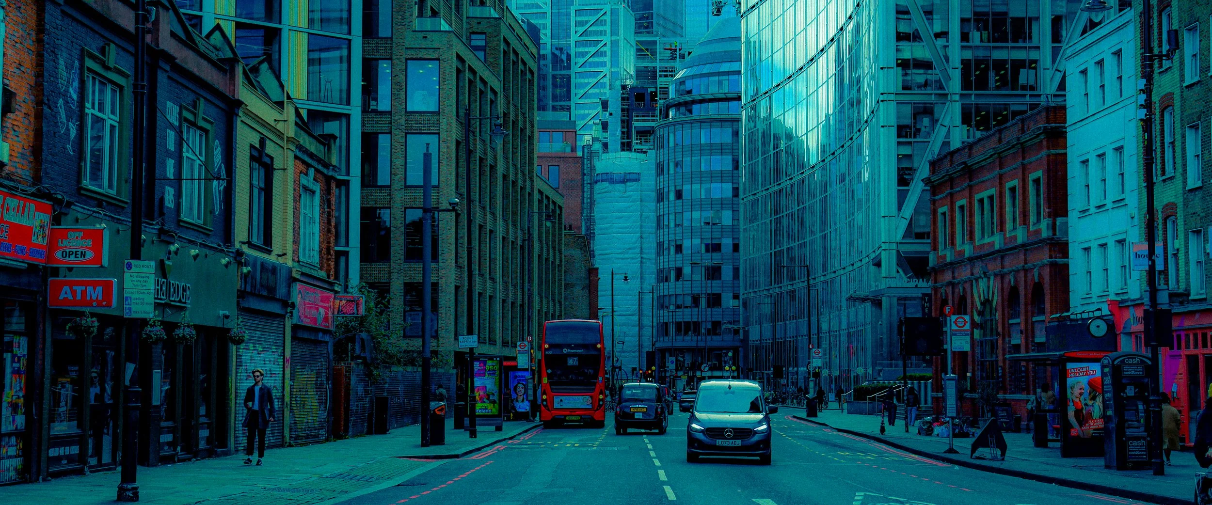 Vista de una calle en una ciudad moderna con edificios altos de vidrios y algunos de ladrillos, autobús doble piso rojo, vehículos y personas caminando.