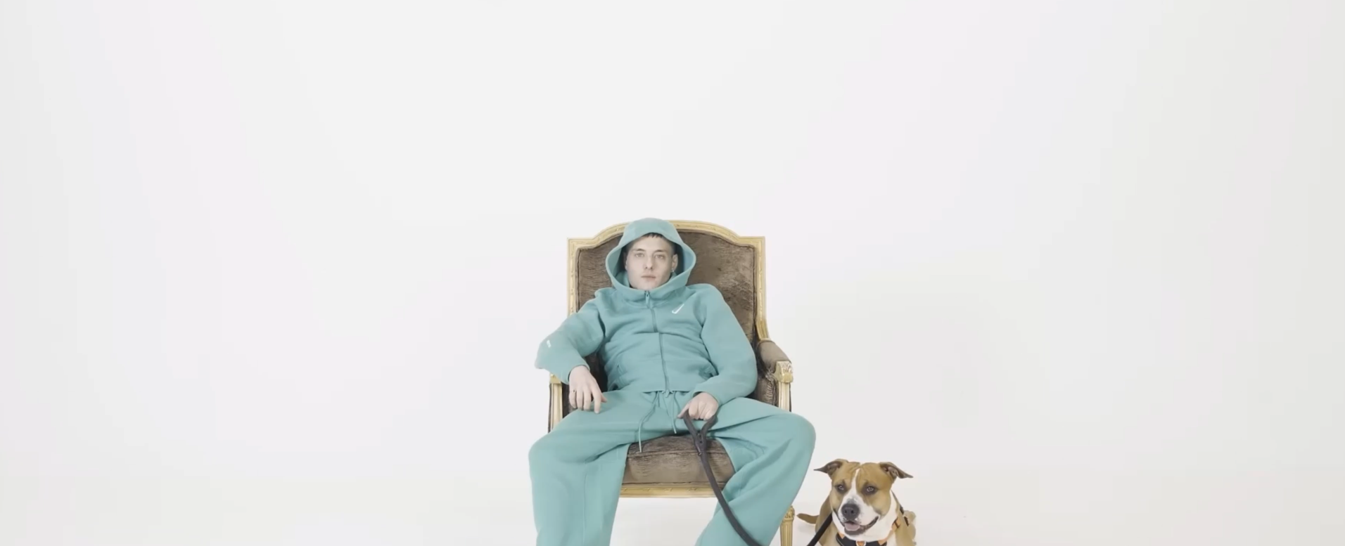 Una persona joven en ropa deportiva azul claro, sentada en una silla antigua, acompañada de un perro de raza pitbull con collar, en un fondo blanco y sin otros elementos visibles.