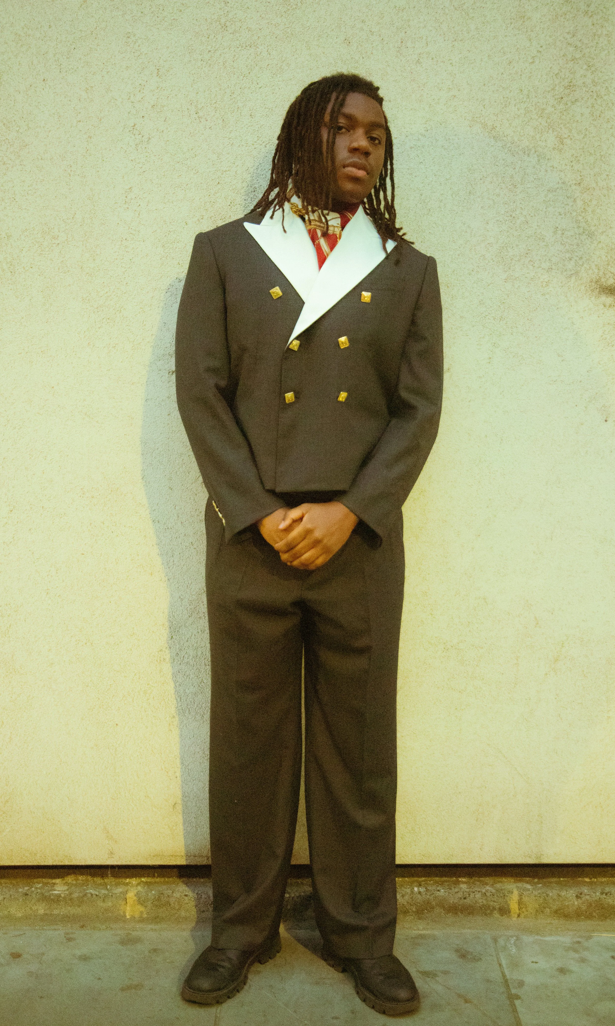 Joven vestido con traje formal oscuro y detalles dorados, de pie contra una pared clara con expresión seria.