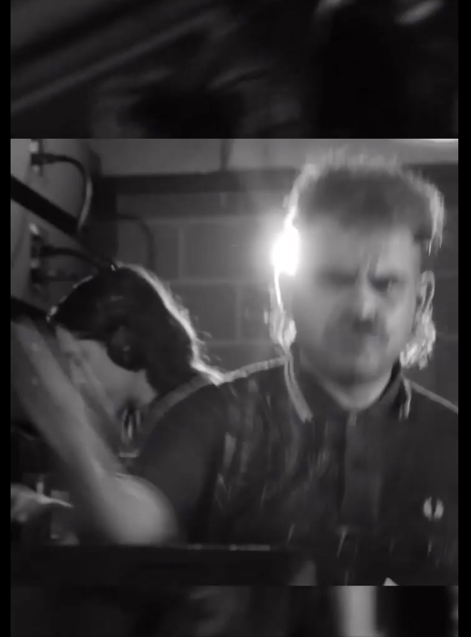 Joven con cabello largo y levantado, con expresión facial seria, en un ambiente con iluminación tenue y fondo de ladrillos, acompañado por una persona de cabello oscuro y largo, en un espacio que parece una bodega o taller.