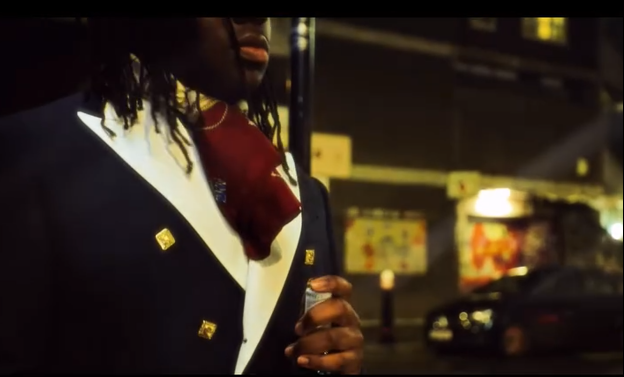 Primer plano de un hombre con chaqueta formal negra y pañuelo rojo, en un entorno urbano nocturno con autos y edificios iluminados.