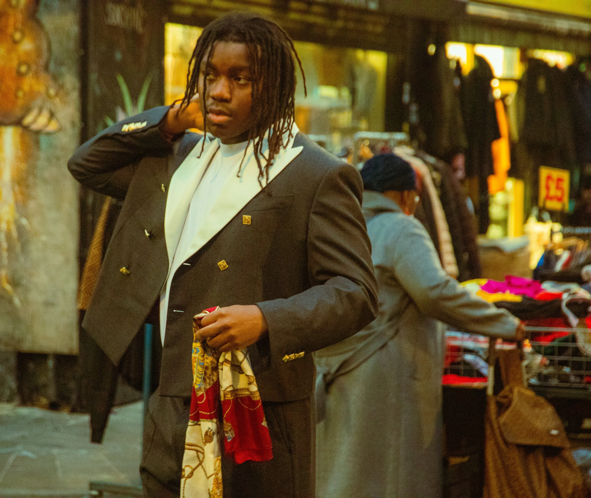 Joven en un mercado al aire libre ajustando su saco y sosteniendo una bufanda colorida.