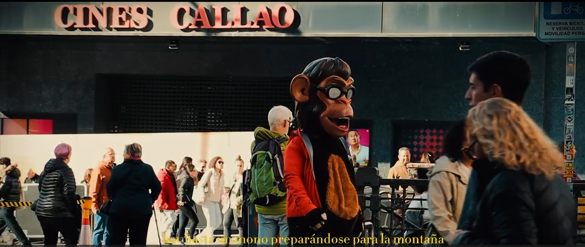 Escena en la entrada de un cine llamado 'Cines Callao', con personas caminando y un personaje disfrazado de mono con gafas en primer plano, en medio de un ambiente urbano y concurrido.