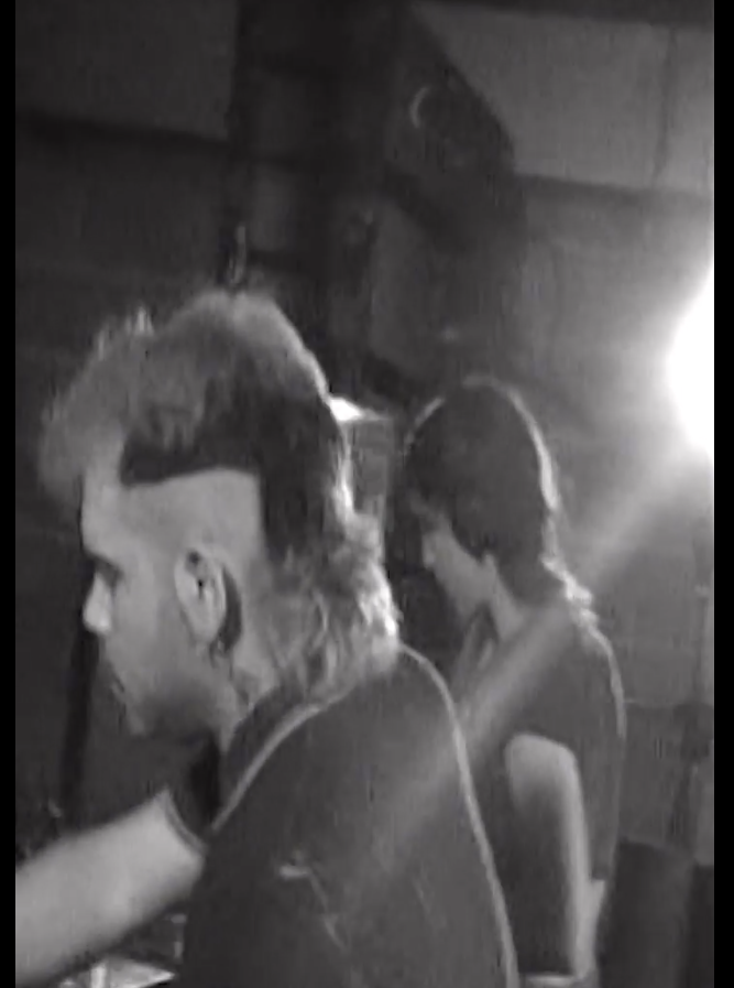 Dos personas en un ambiente oscuro con luz intensa en el fondo, parecen estar concentradas en alguna tarea.