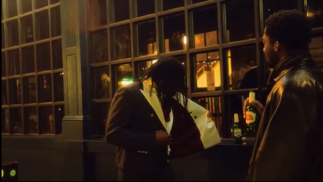 Dos personas bebiendo en un bar, una con cabello rasta y un saco negro, la otra con barba y chaqueta de cuero.