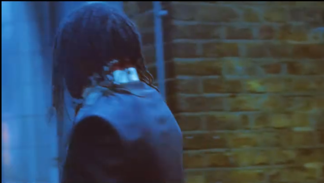 Persona con cabello largo y oscuro caminando por una calle, vista de espaldas, con pared de ladrillos en el fondo.