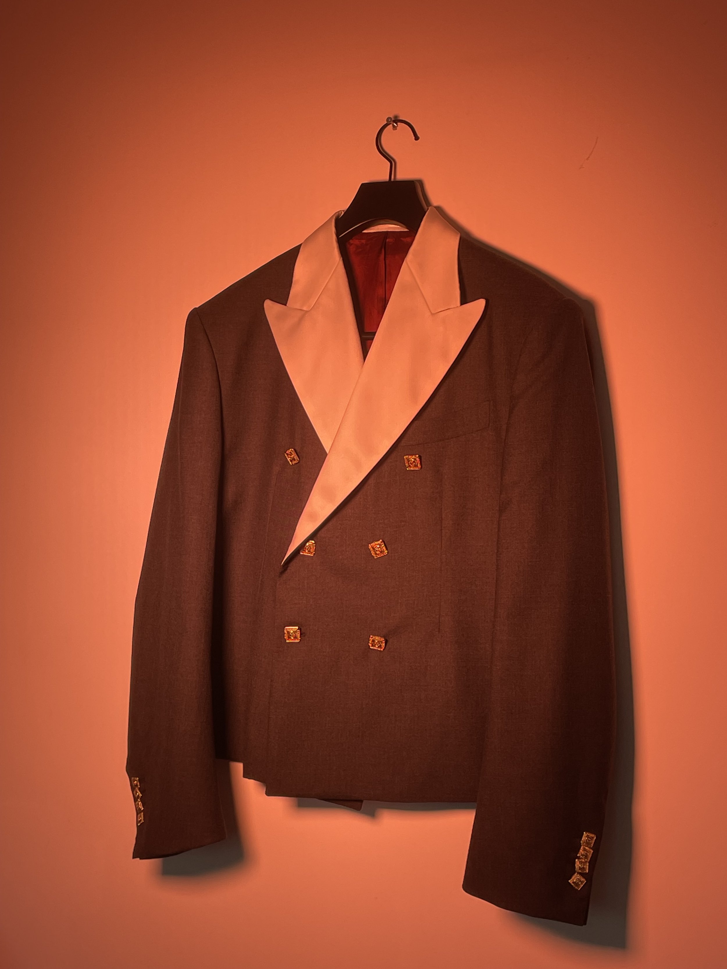 Traje de chaqueta doble botonadura de color gris oscuro con solapa y forro beige, colgado en una percha sobre una pared con iluminación cálida.