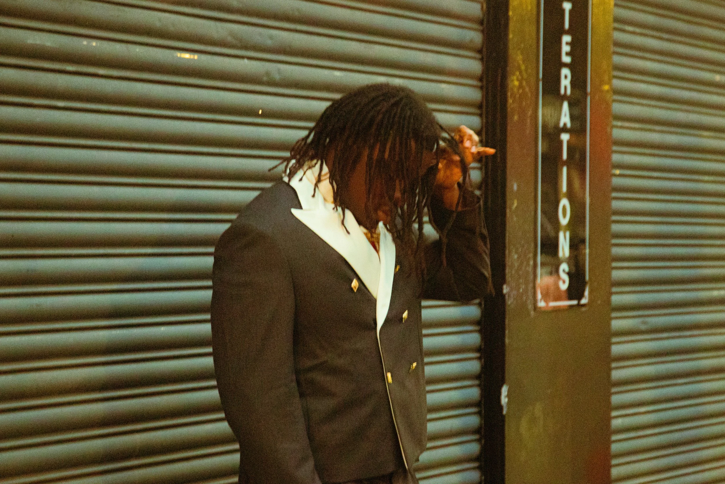 Persona con cabello rizado y gafas, vestida con un saco negro con detalles dorados, apoyado en una columna en un área con persianas metálicas y un letrero que dice 'TERRATONS'.