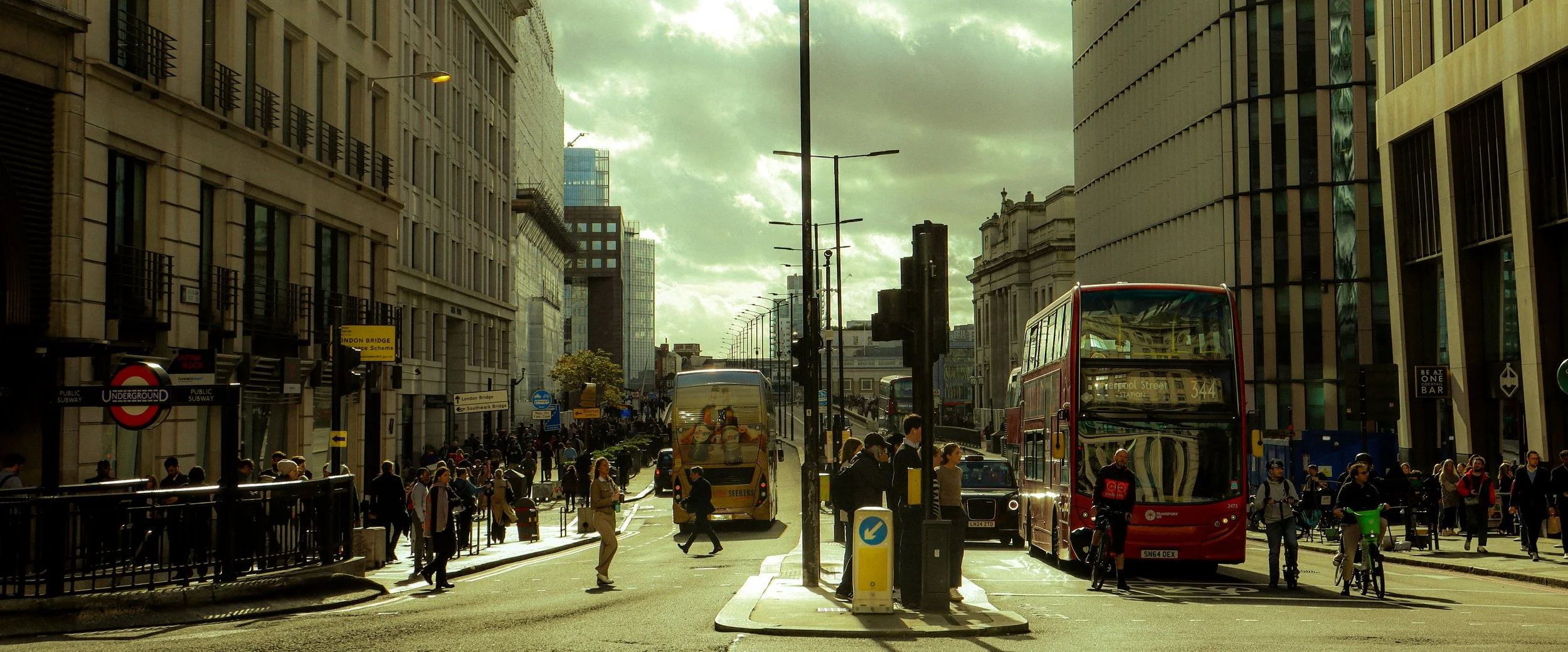 Vista de una calle concurrida en Londres con autobuses rojos, personas caminando y edificios altos a ambos lados, bajo un cielo con nubes.