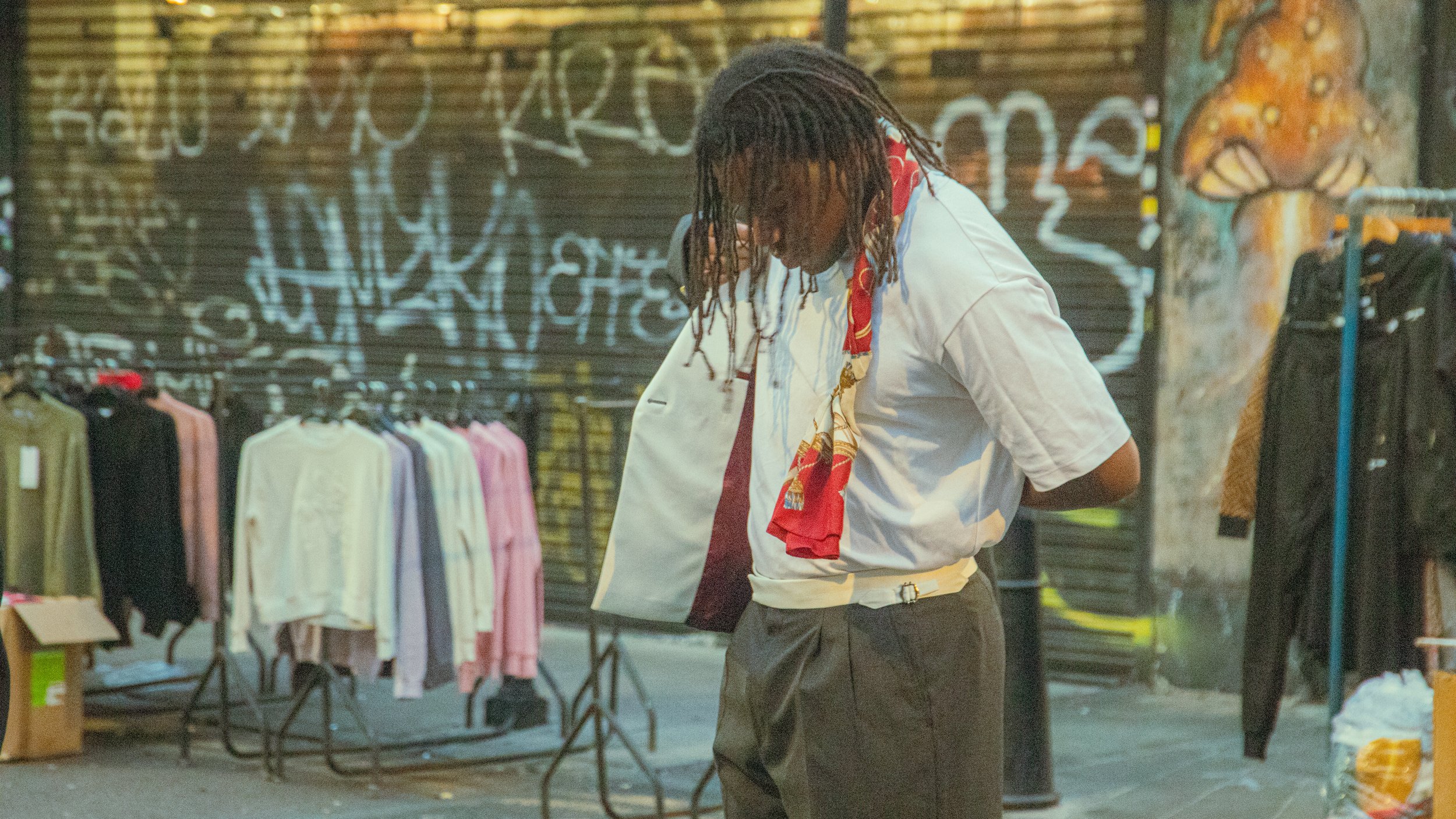 Persona vestida con camiseta blanca, pantalón oscuro y bufanda roja, parada en una calle con puestos de ropa, con letras y graffitis en una pared de fondo.