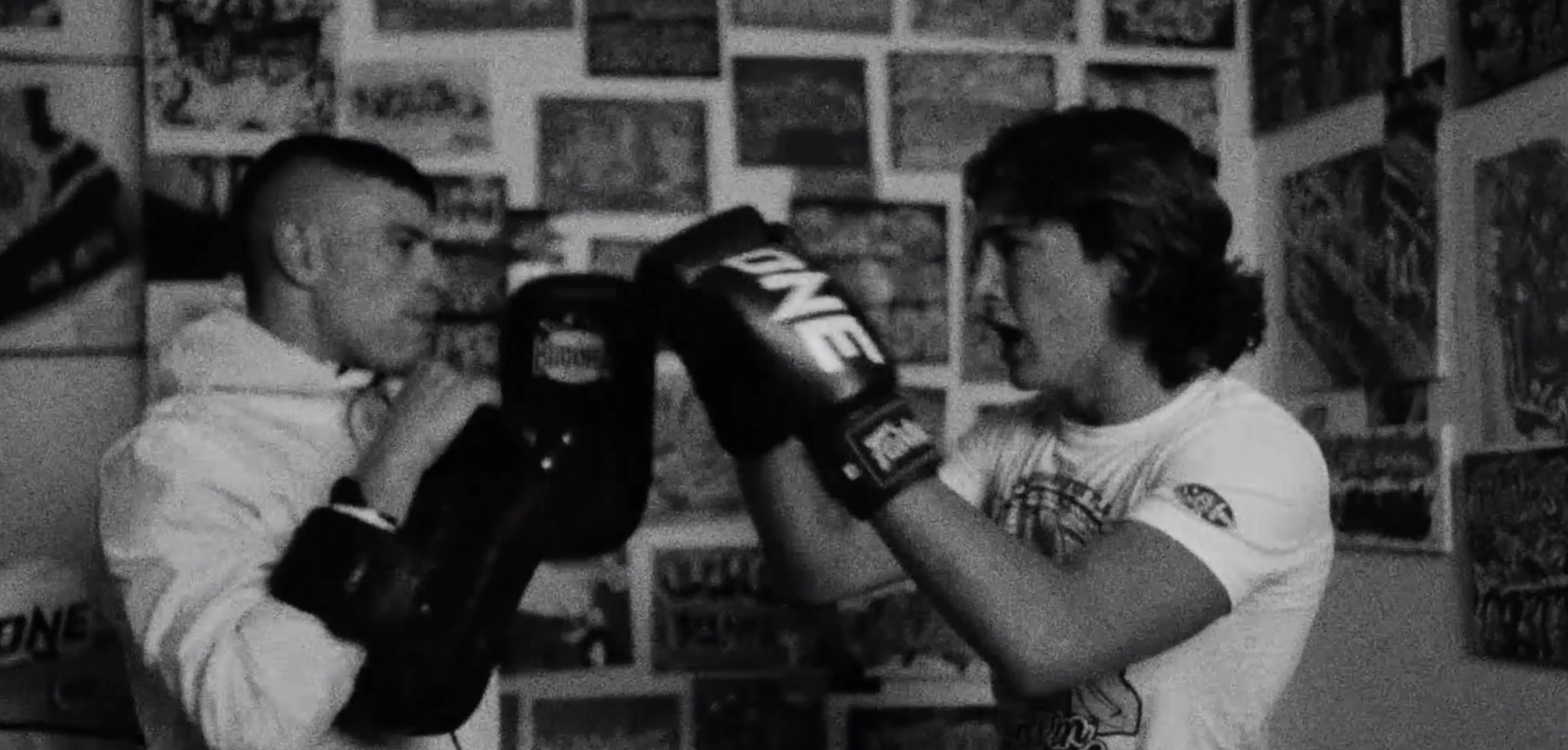 Dos niños entrenando en un ring de boxeo en una habitación decorada con posters en las paredes.