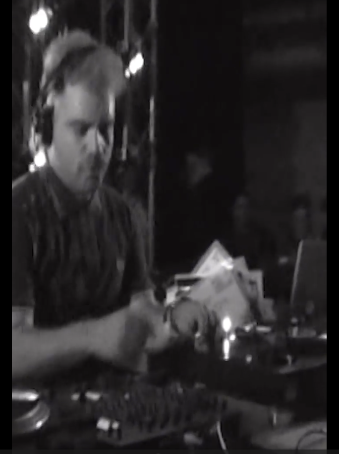 Persona mezclando música en una cabina de DJ en un ambiente oscuro.