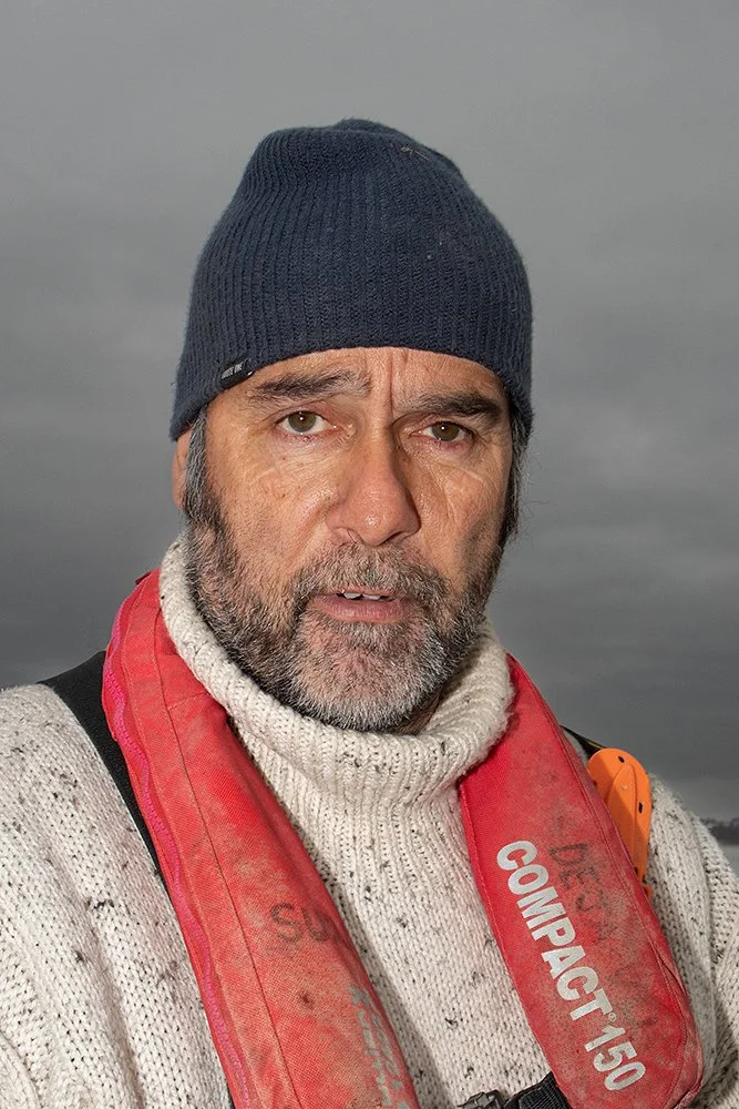 A man with a gray beard wearing a navy blue beanie, a cream-colored sweater, and a red life jacket outdoors on a cloudy day.