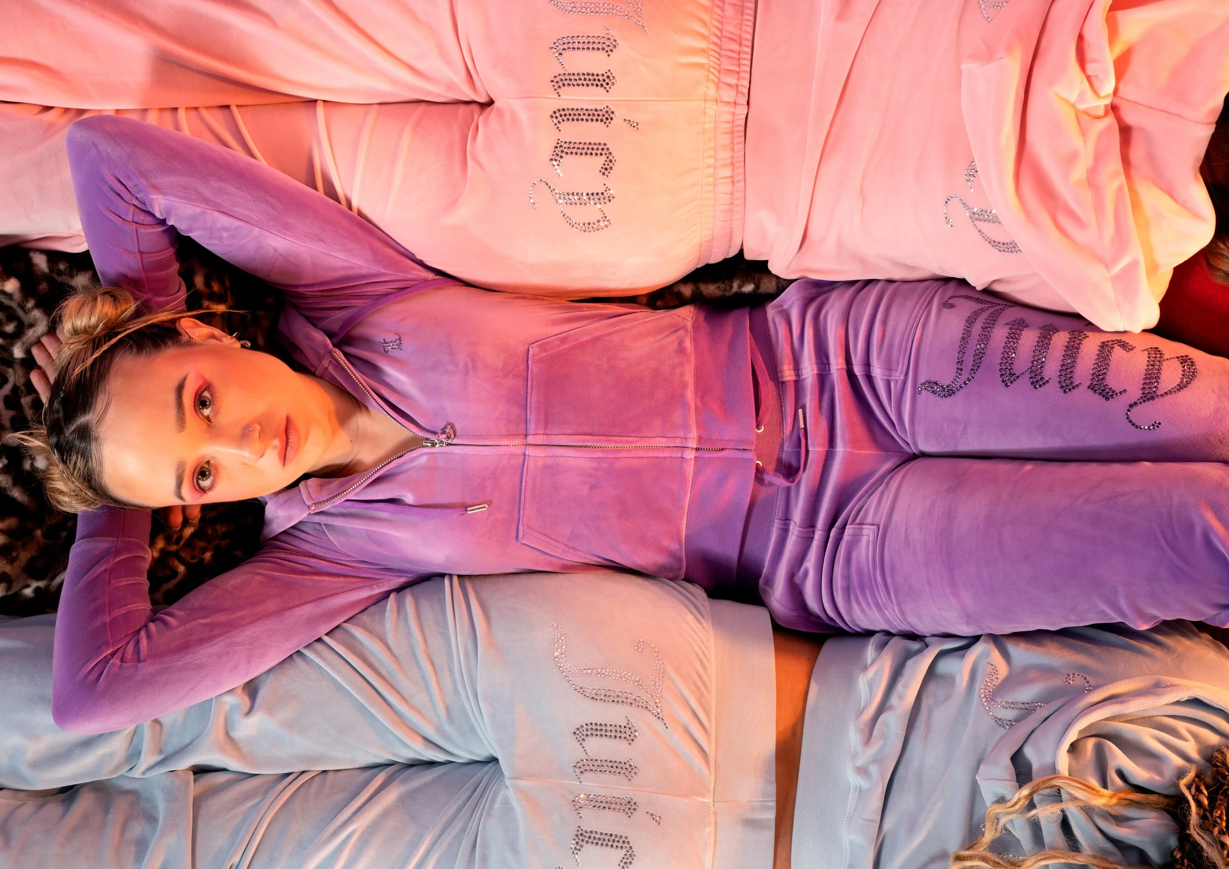 Woman lying on her back on a leopard print rug, wearing a purple velour tracksuit with rhinestone embellishments, surrounded by pastel-colored hoodies with similar rhinestone designs.