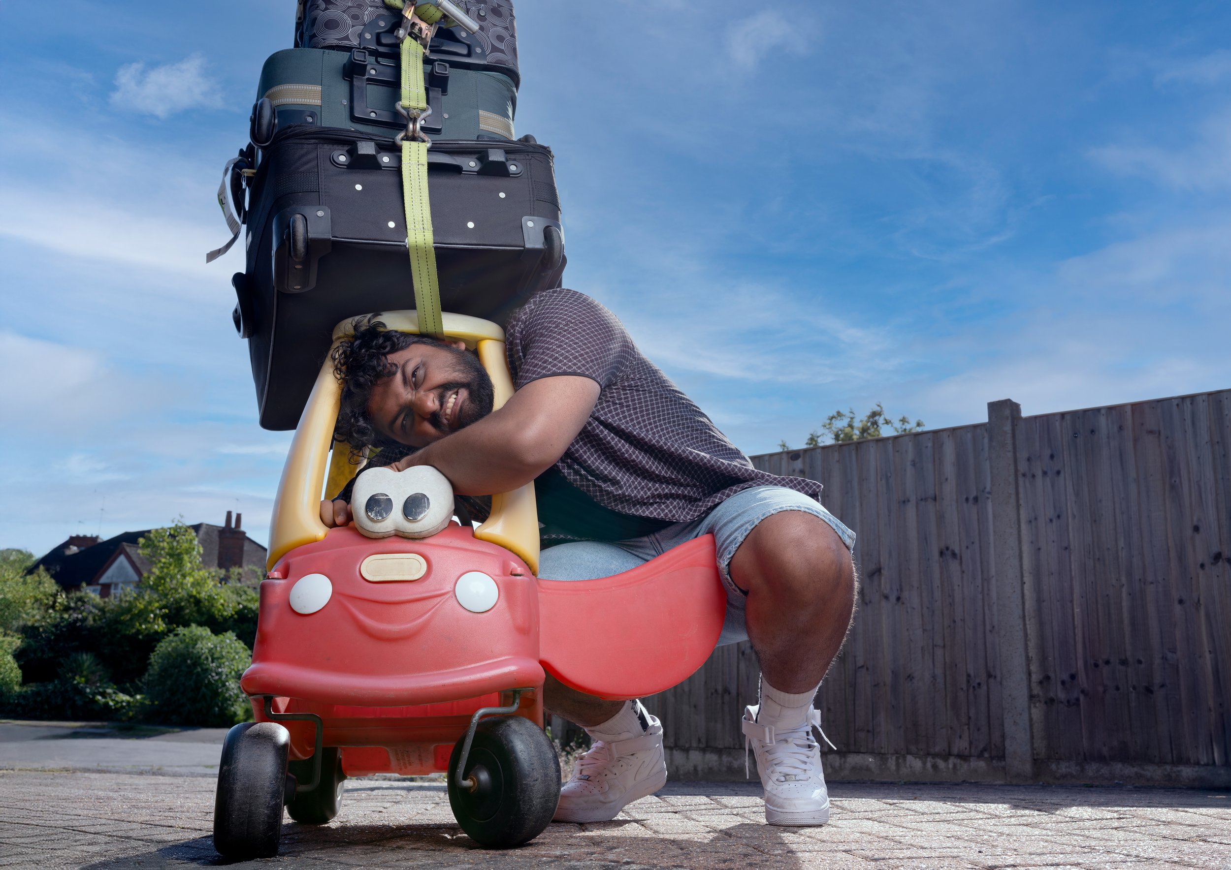 A young man with curly hair and a beard crouches next to a red children's toy car with a smiling face design. He is hugging the car's steering wheel while smiling at the camera. The man is wearing a short-sleeved checked shirt, denim shorts, and whit