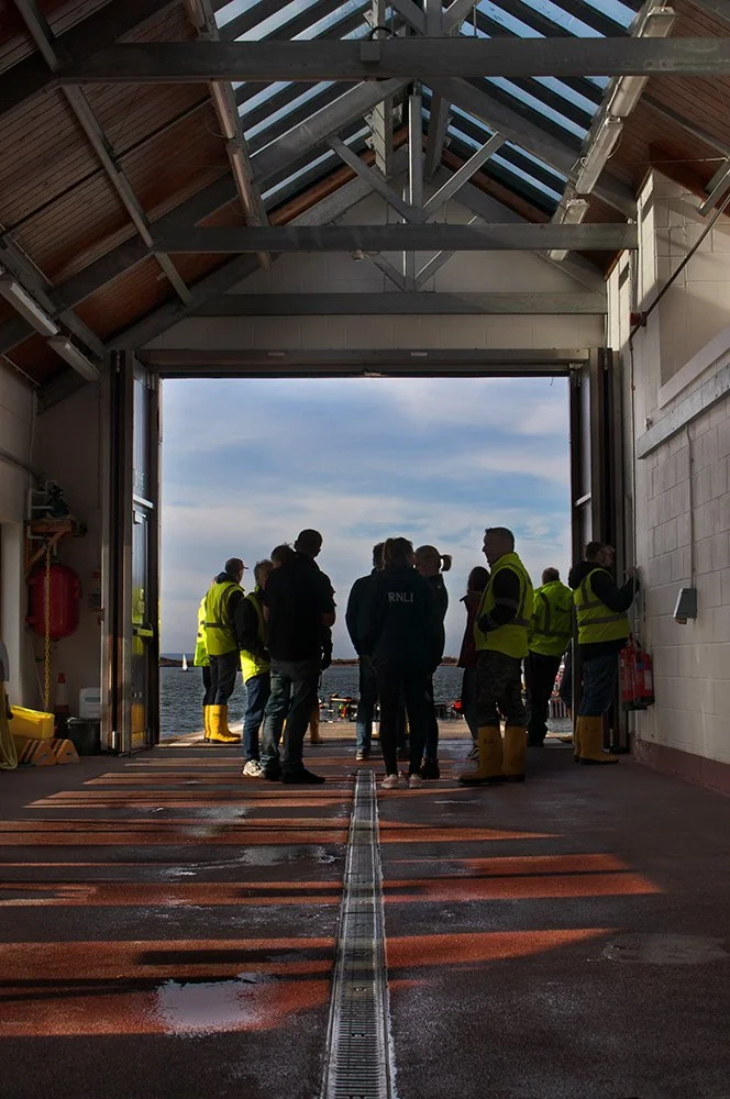 Group of people, some wearing yellow safety vests and boots, standing inside a large ferry or ship, looking out towards the water through an open hatch, with a cloudy sky and water visible in the background.