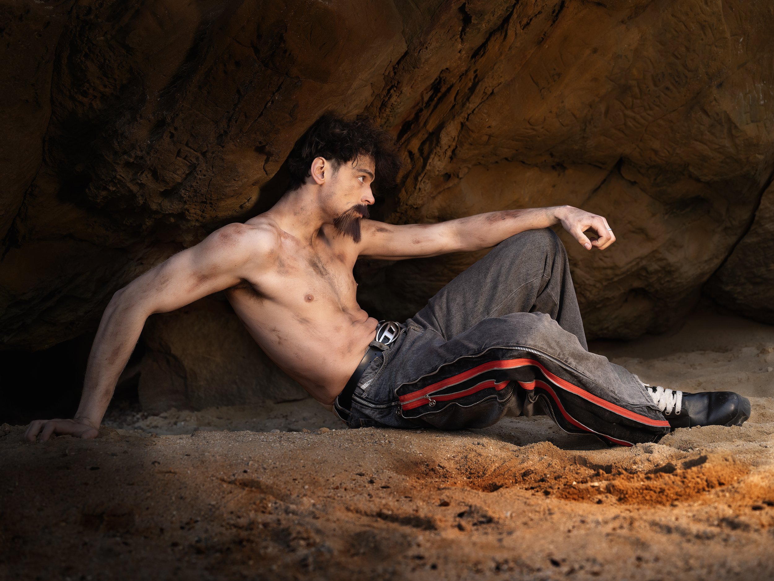 A shirtless man with a beard and disheveled hair, sitting against a large rock formation, in a sandy environment.
