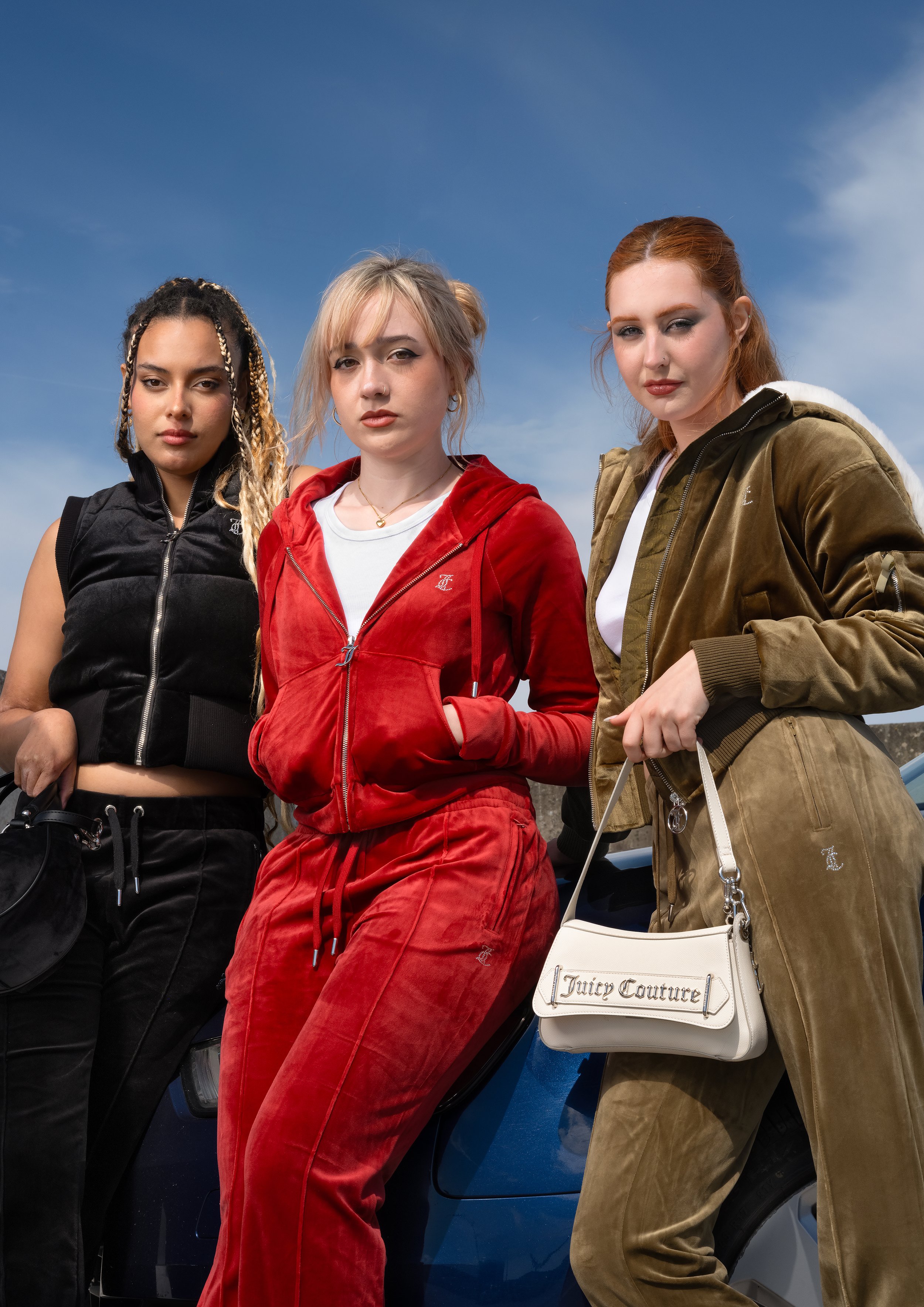 Three women standing outdoors near a blue car, dressed in casual sporty outfits, under a clear blue sky.