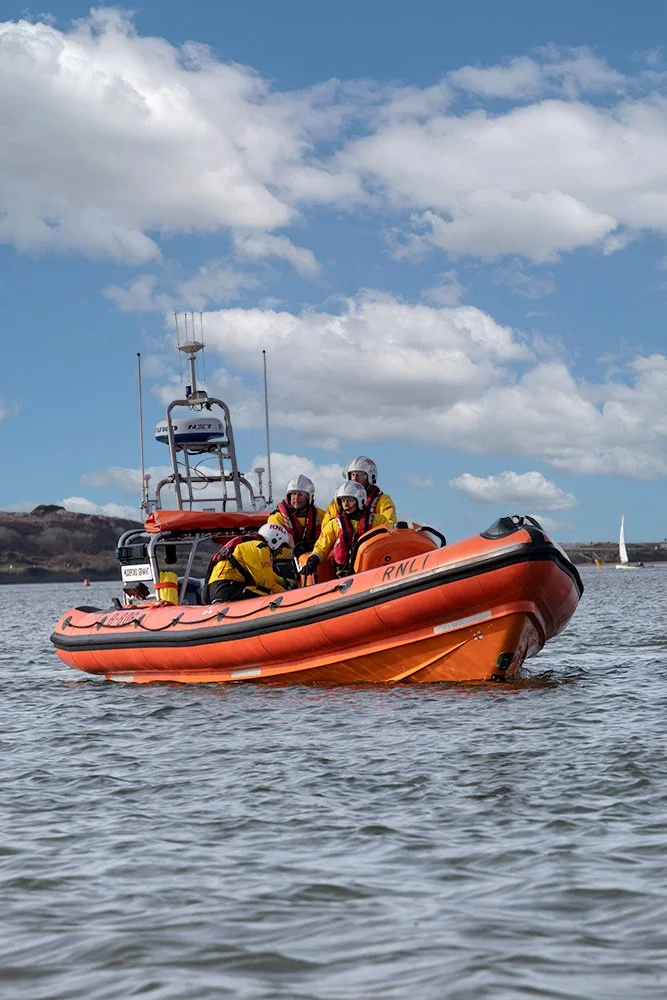 Rescue team in yellow and red uniforms on an orange rigid inflatable boat on the water, with a blue sky and scattered clouds in the background.