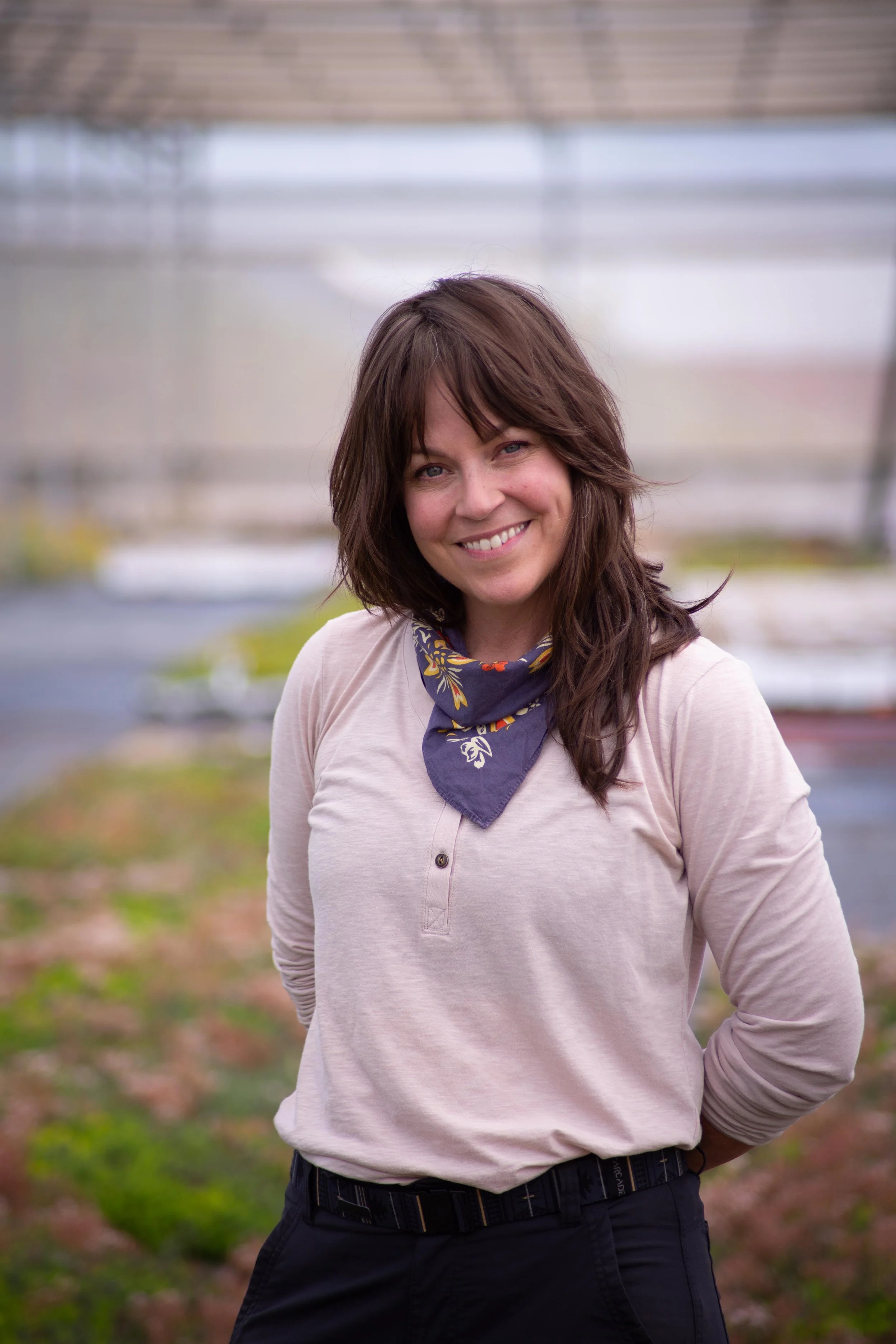A woman smiling outdoors with a background of greenhouse structures and a dirt and grass ground.