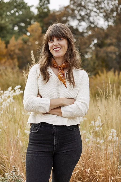 Smiling woman with brown hair and bangs, wearing a cream sweater, black jeans, and an orange patterned scarf, standing in a field with autumn trees in the background.