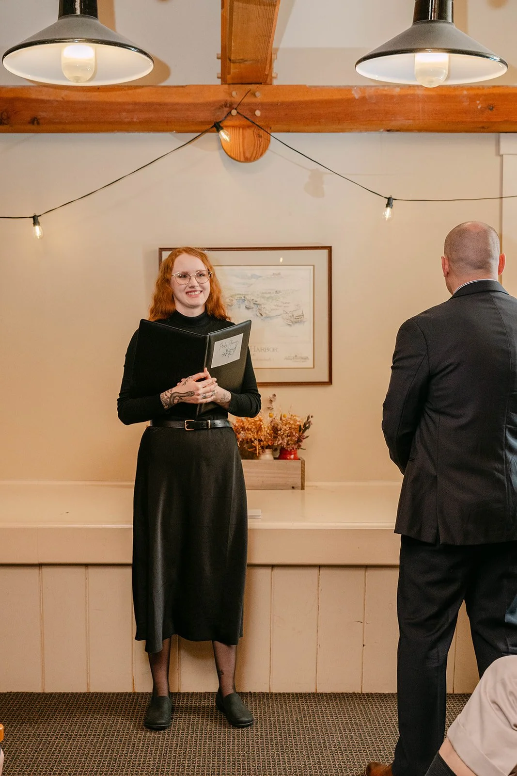 A woman with red hair, glasses, and tattoos wearing a black dress stands holding a folder, smiling during a ceremony. A man in a black suit is facing her. The setting has warm lighting, with wooden beams, a framed picture of a harbor on the wall, and a potted plant with dried flowers behind them.