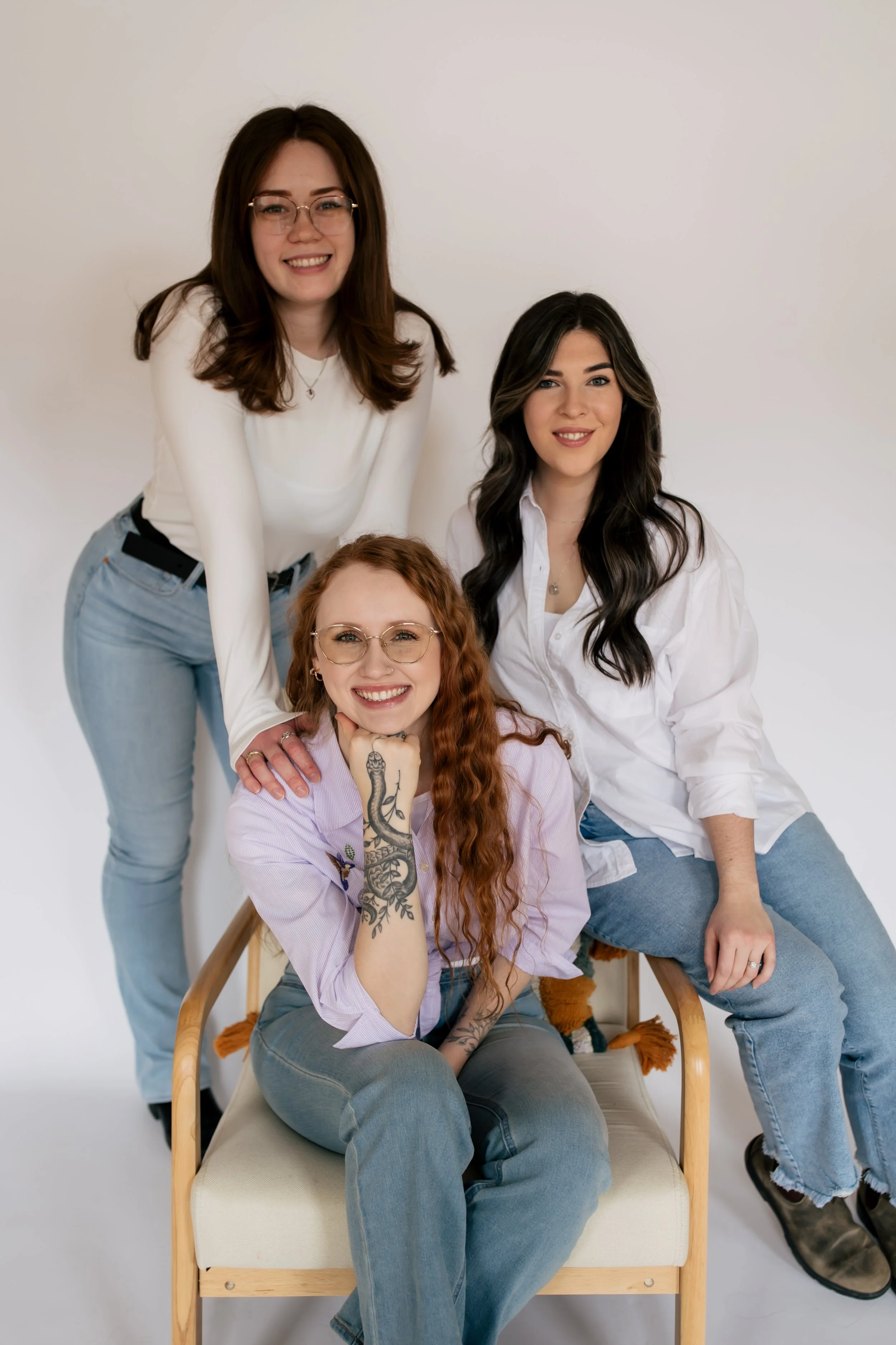 Four women posing together against a plain white background, with one sitting on a chair and the others standing around her.