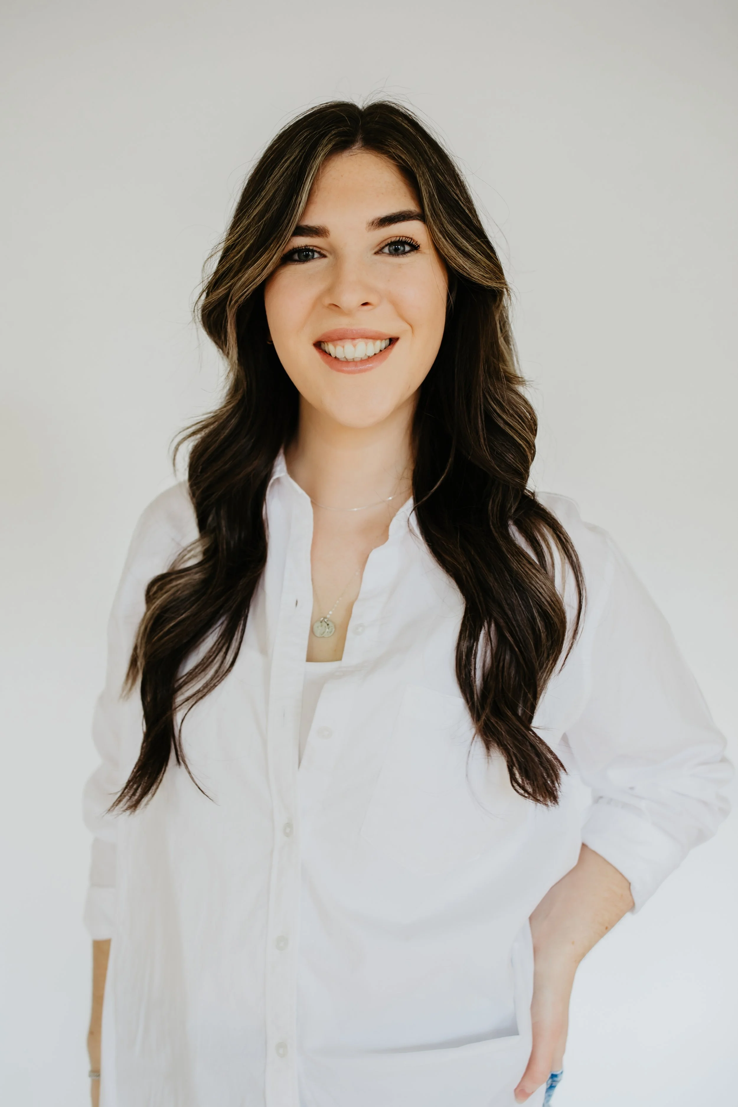 A young woman with long, dark wavy hair wearing a white button-down shirt, smiling at the camera, standing against a plain light background.