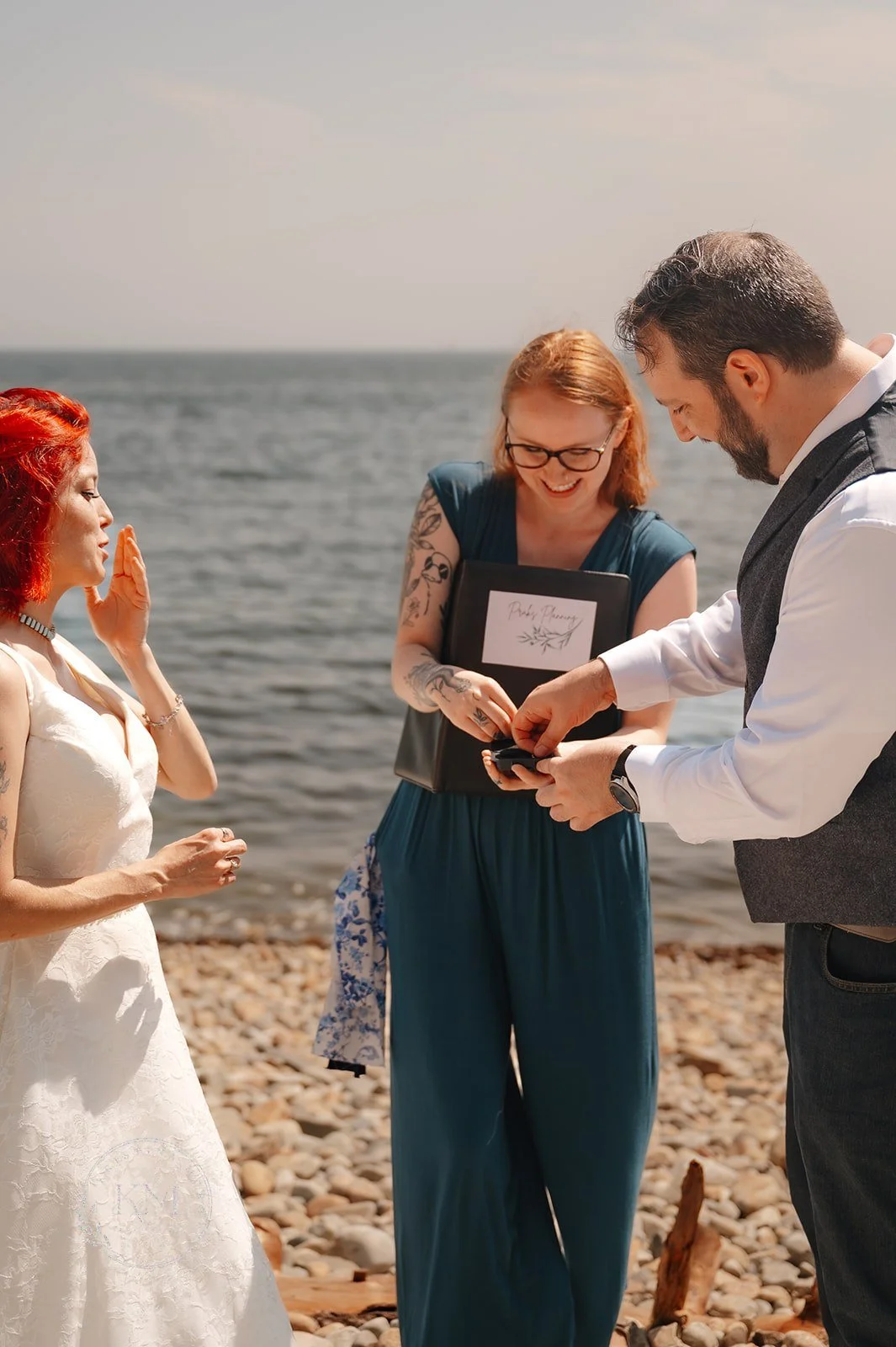A wedding ceremony on a pebbled beach with a woman in a white dress, a woman in blue, and a man in a white shirt and dark vest. The officiant is holding a device, possibly reading vows or conducting the ceremony, while the bride looks on with a smile.