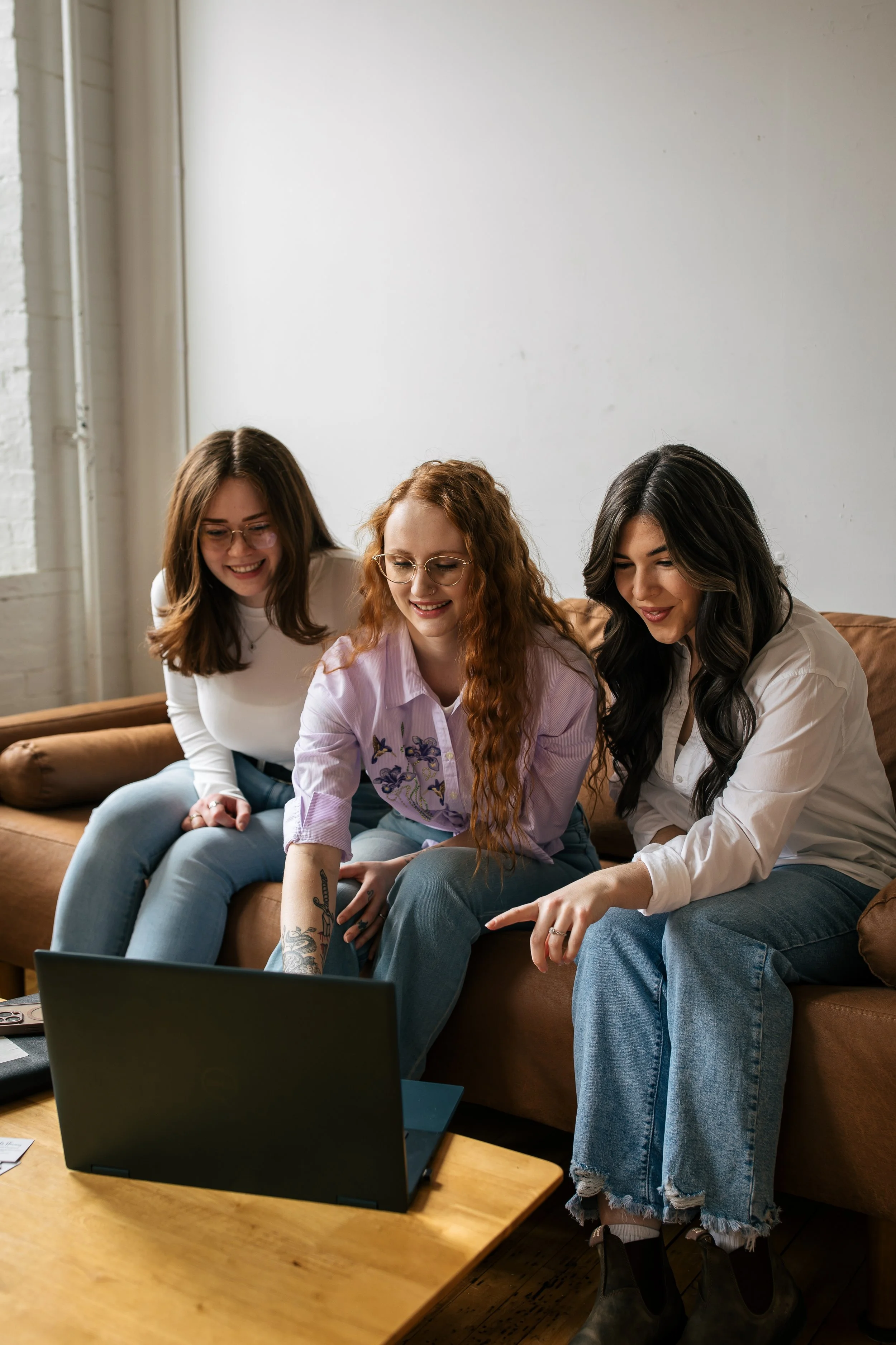 Three women sitting on a brown couch, looking at a laptop, smiling, in a room with a white wall and natural light.