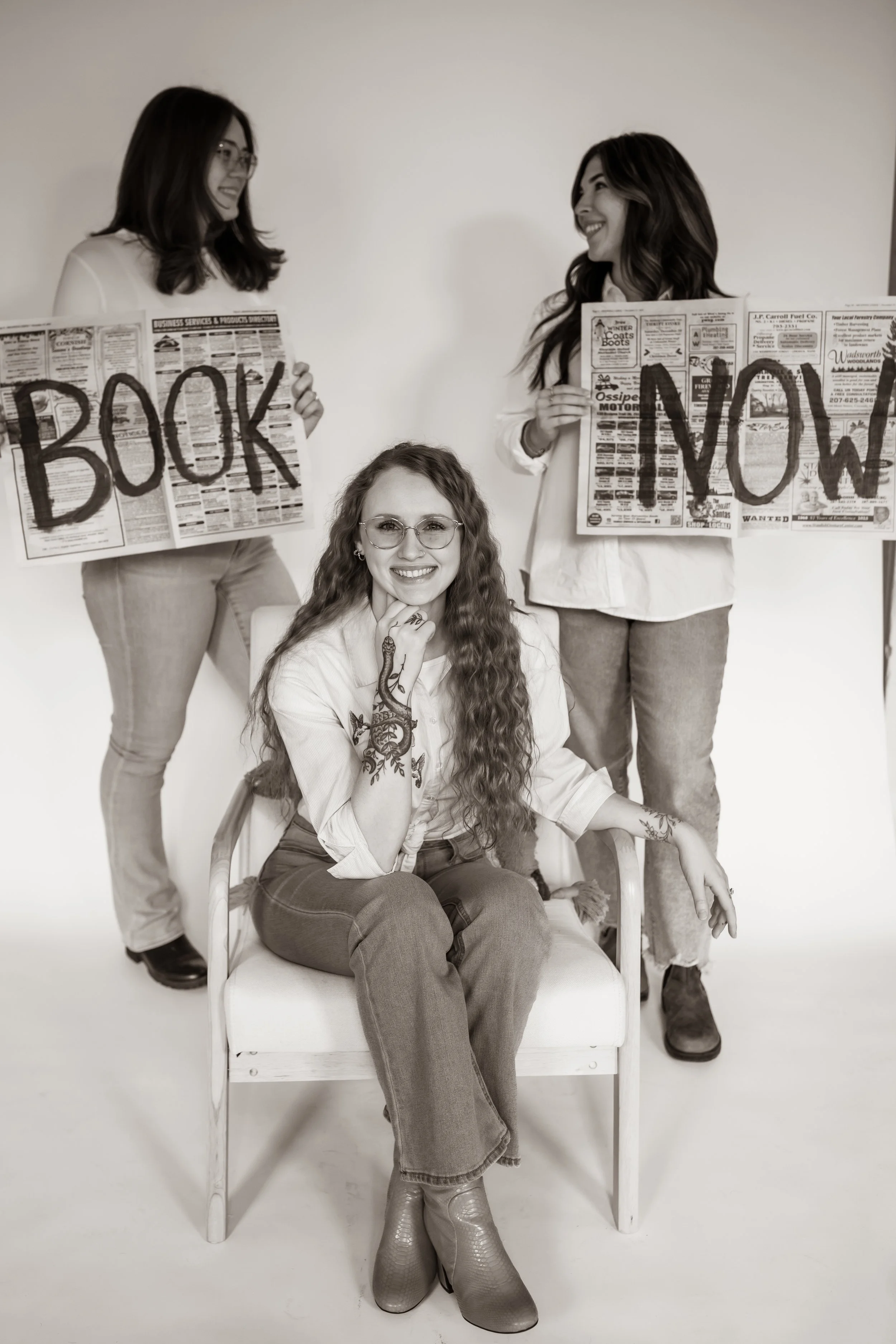 Three women, one sitting in a chair and two standing behind her, holding signs that spell "BOOK" and "NOW". The woman in the chair has long curly hair, glasses, tattoos, and is smiling at the camera.