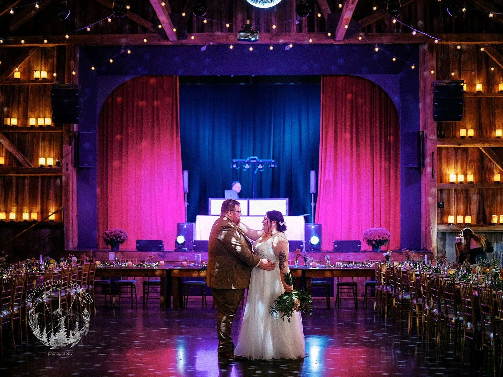 A couple on their wedding dance in a rustic wooden barn with tables and chairs decorated for a celebration, with warm lighting and pink floral arrangements in the background.