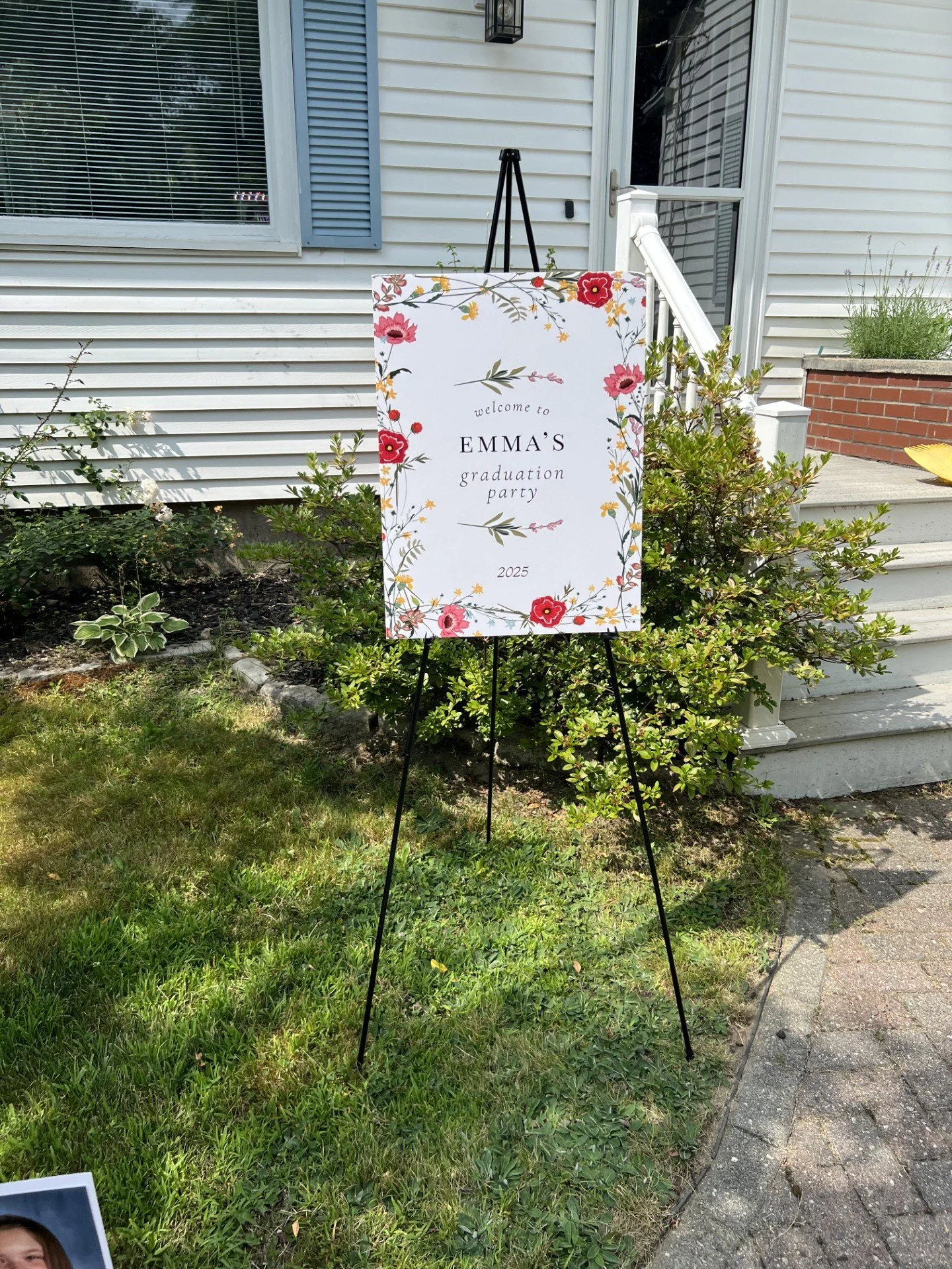 A floral welcome sign on a black easel outside a house with white siding and stairs, inviting guests to Emma's graduation party in 2025.