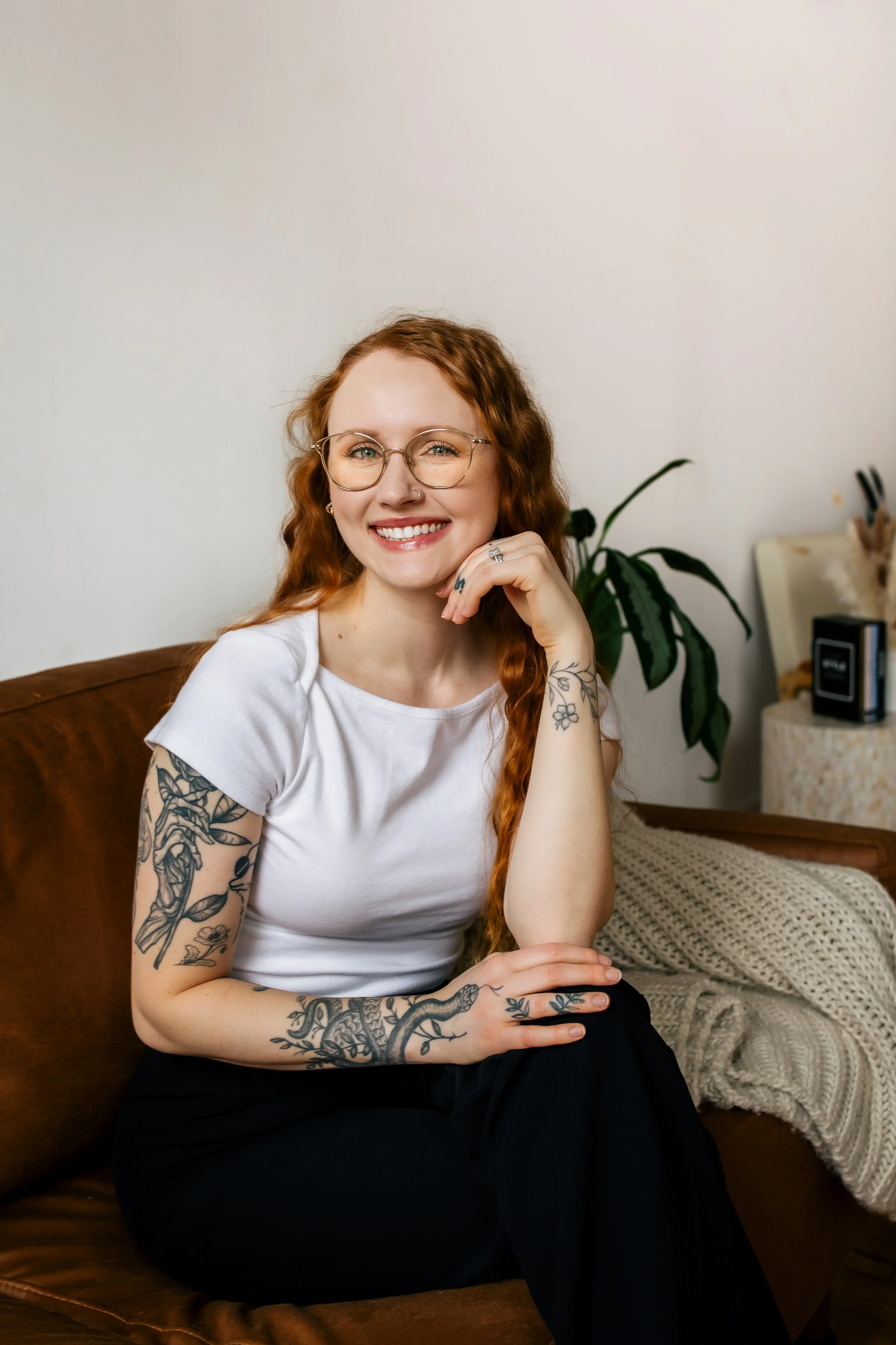 A smiling woman with long red hair, tattoos on her arms, wearing glasses and a white T-shirt, sitting on a brown sofa in a cozy room with a plant and a blanket in the background.