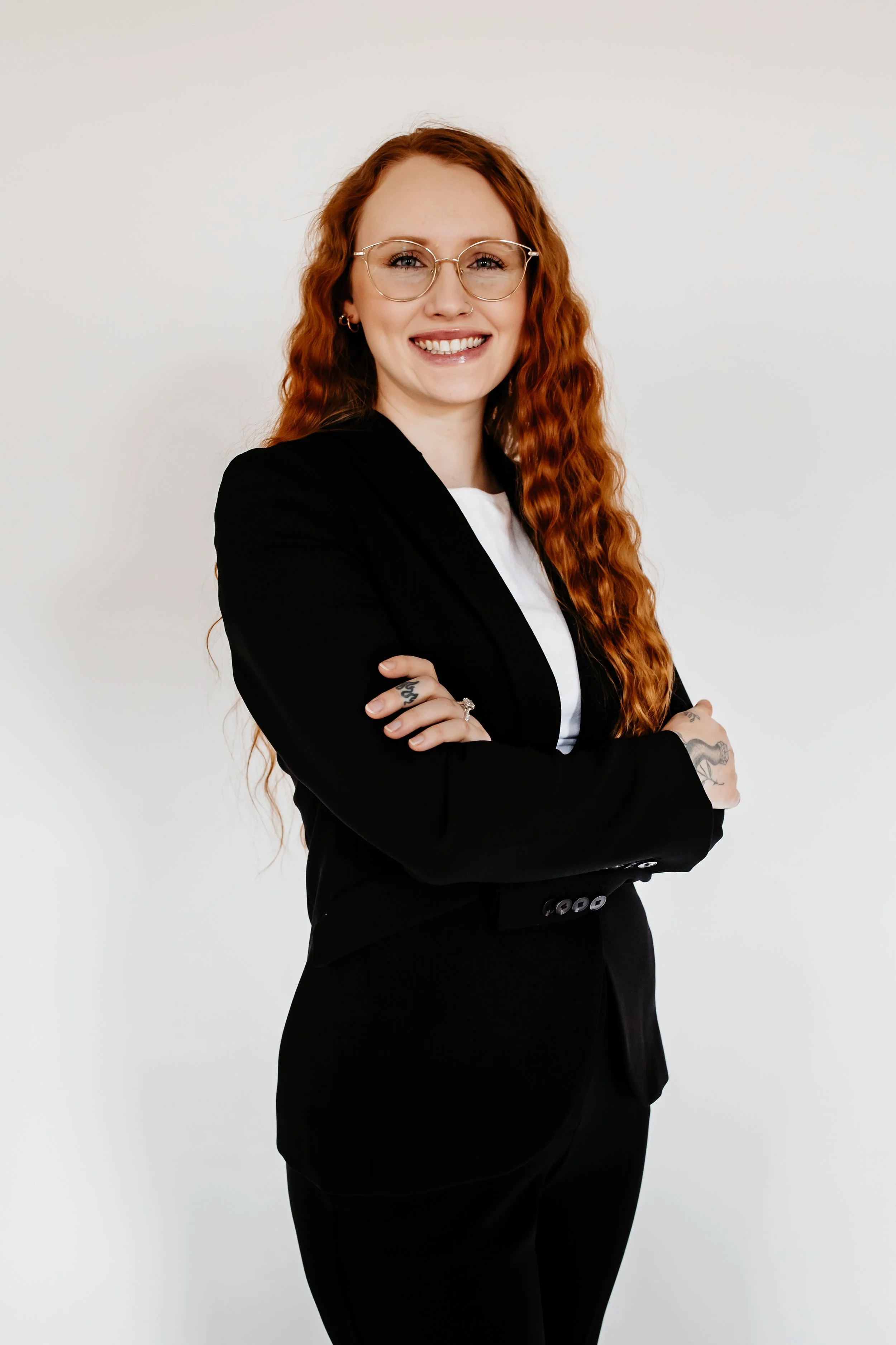 A woman with long curly red hair, glasses, and tattoos on her hands, wearing a black blazer over a white shirt, standing with arms crossed and smiling against a plain white background.