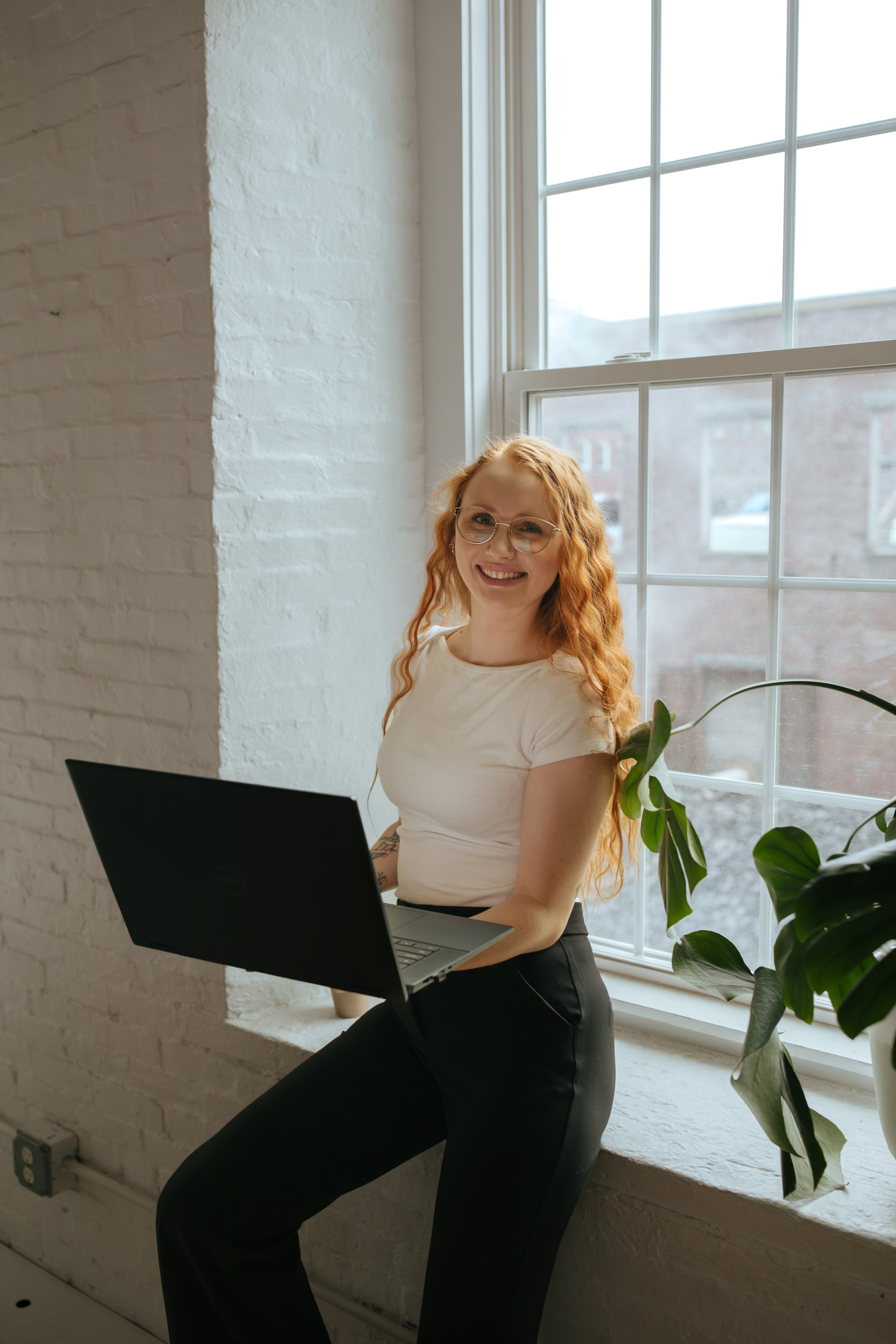 A smiling red-haired woman sitting on a window ledge with a laptop in a bright, modern room with white brick walls and large window.